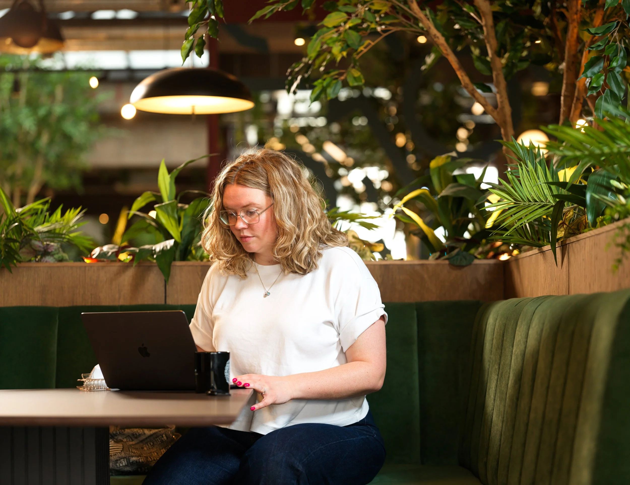 Woman with curly blonde hair and glasses working on a laptop at a table in a lush, green indoor space with hanging lights.