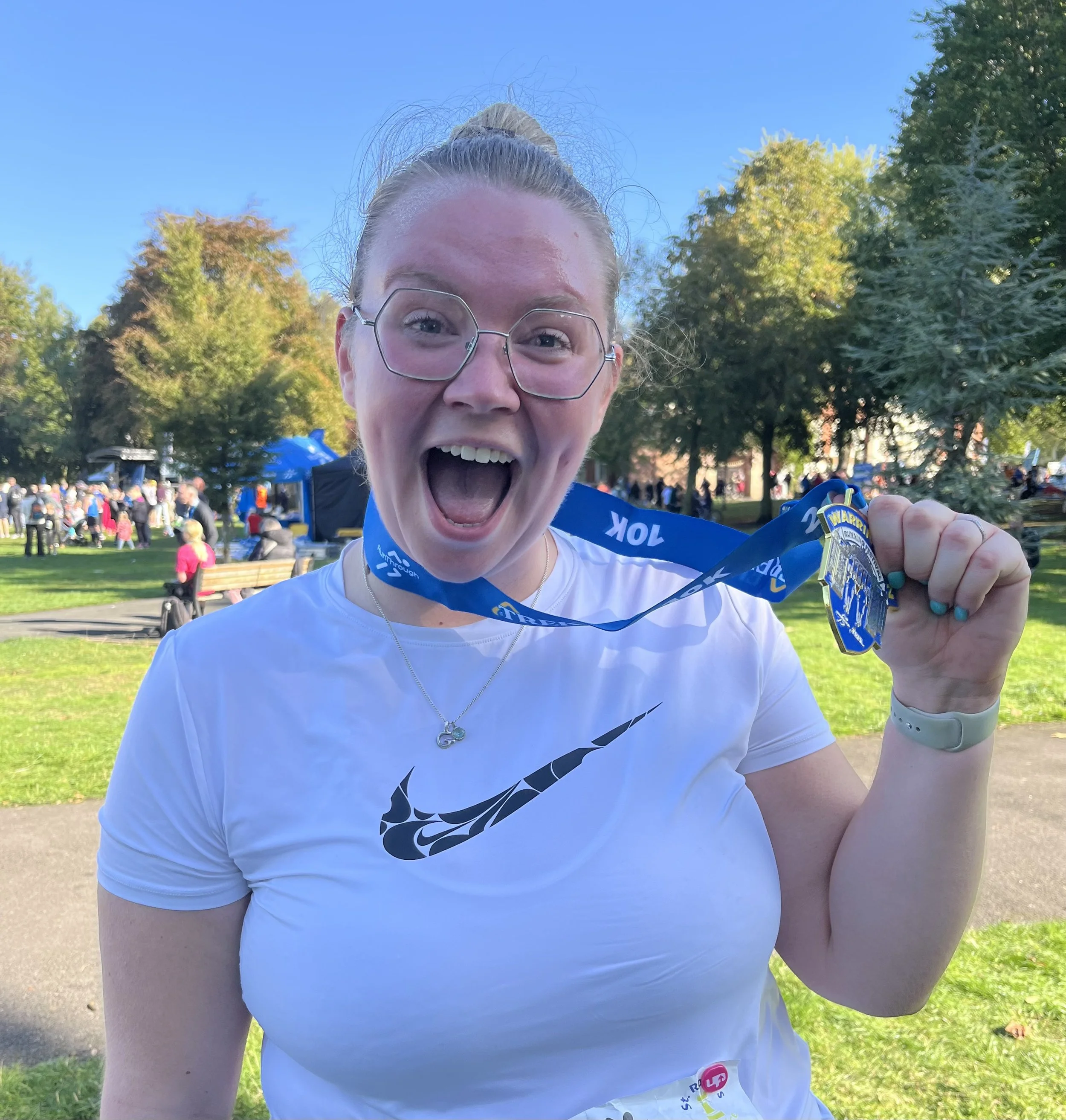 Close-up of a woman with glasses smiling and holding up a medal after a race or event outdoors in a park with trees and people in the background.