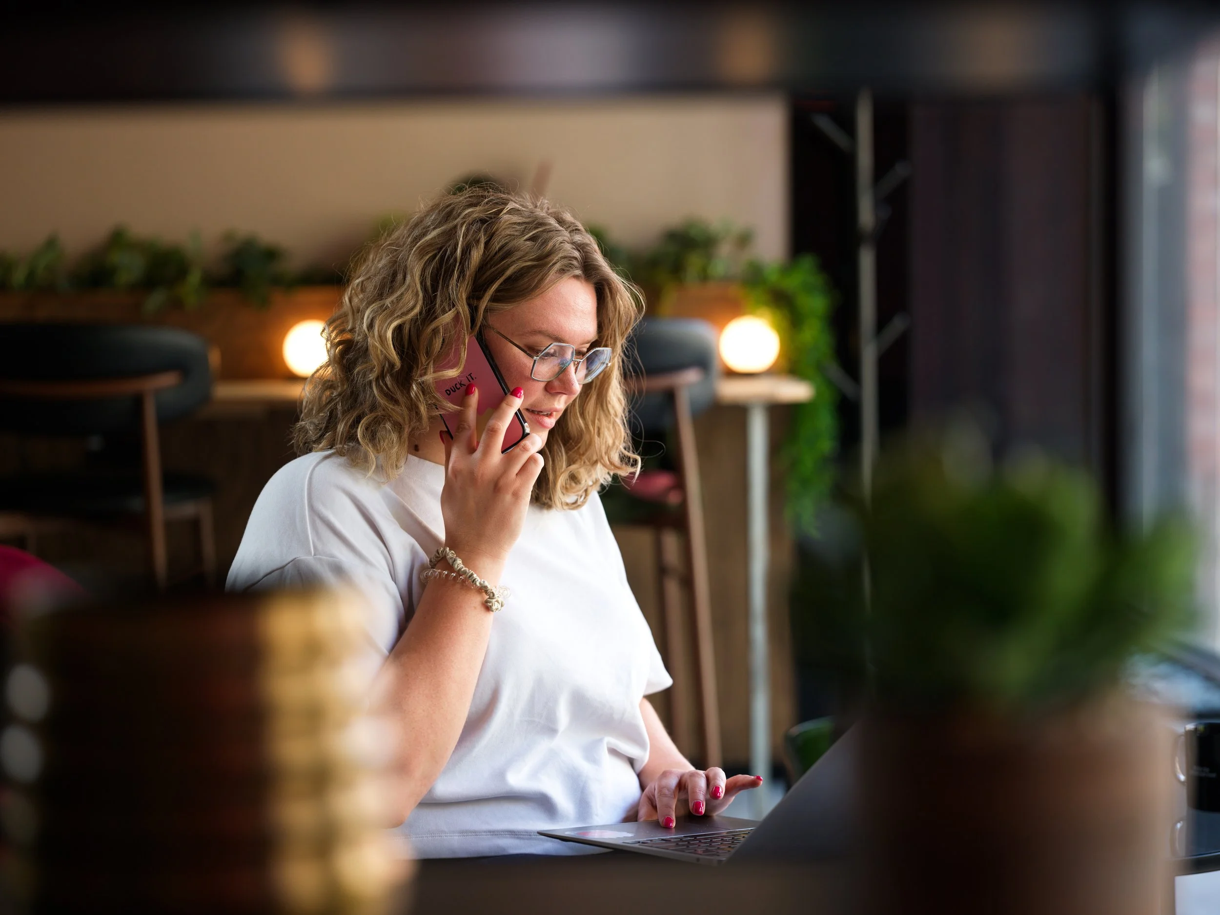 Woman with curly blonde hair and glasses talking on a pink smartphone in a cozy cafe, working on her laptop, with blurred wooden furniture and green plants in the background.