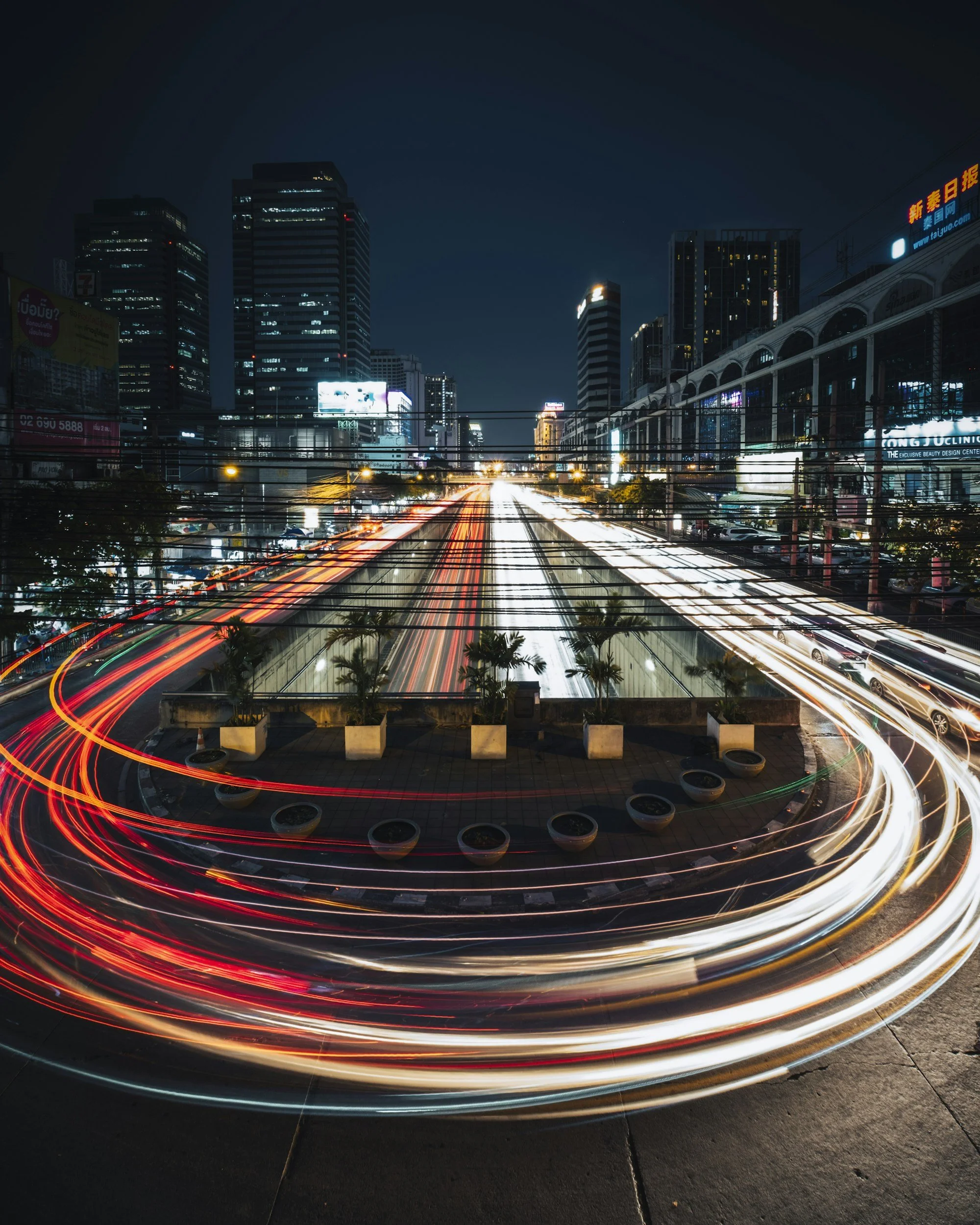 Long exposure photo of light trails from moving vehicles on a city street at night, with tall buildings and billboards in the background.
