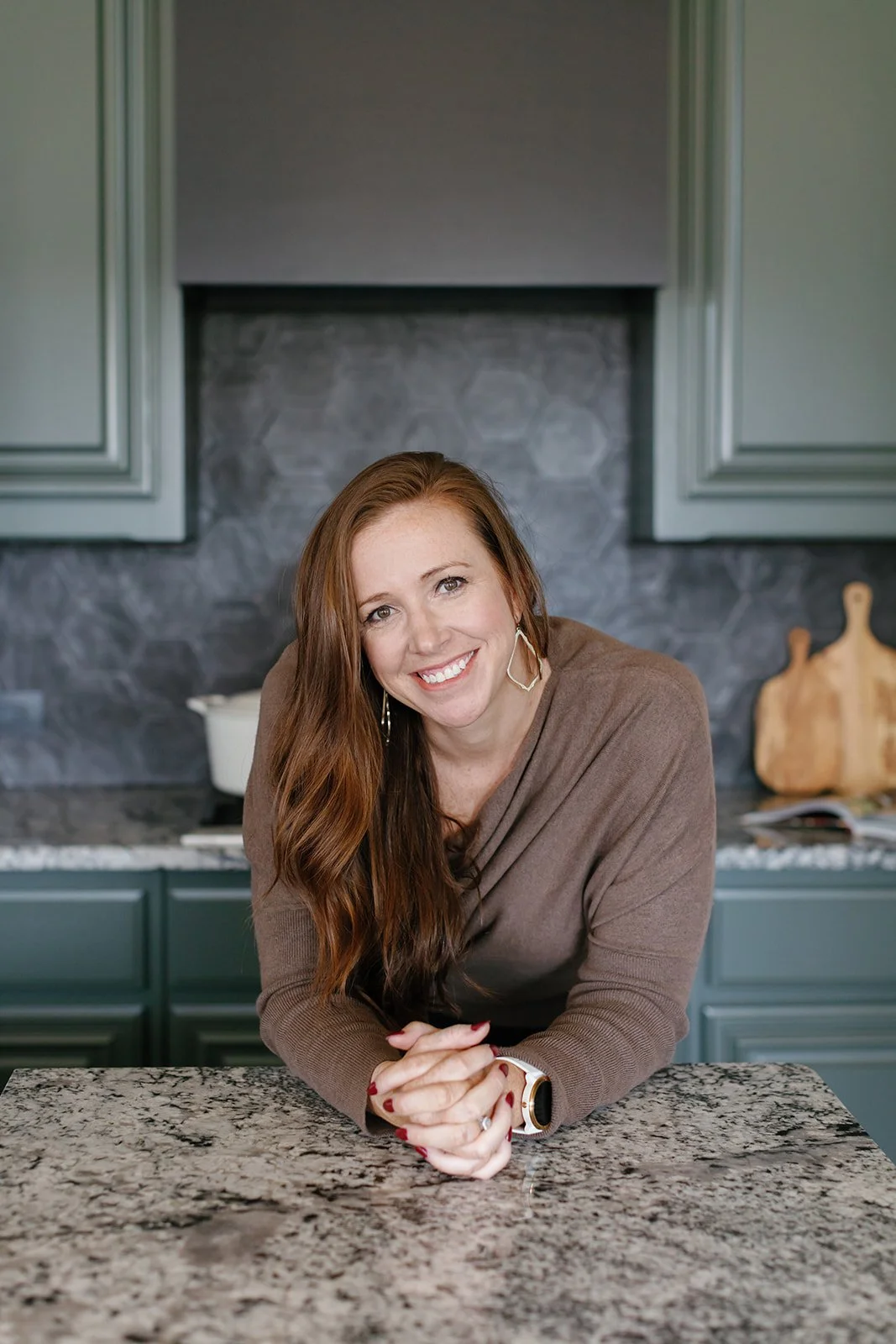 A woman with long red hair and a brown top leaning on a kitchen counter, smiling at the camera.