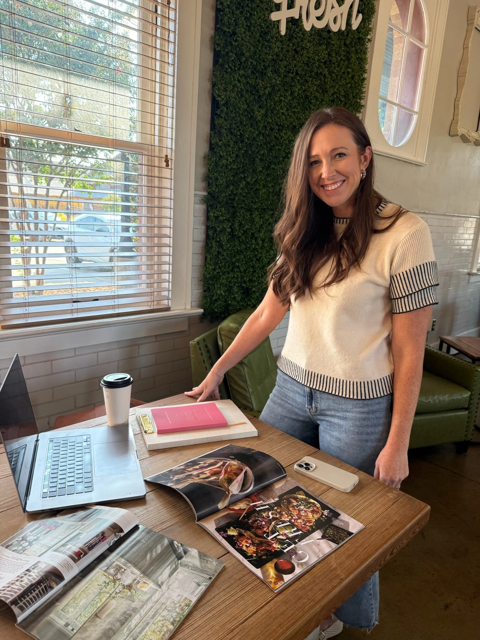 A woman with long brown hair smiling and wearing a cream-colored sweater with black trim, standing at a wooden table with magazines, a laptop, a smartphone, a coffee cup, and other items, in a cozy cafe with a green wall and a window with wooden blinds.