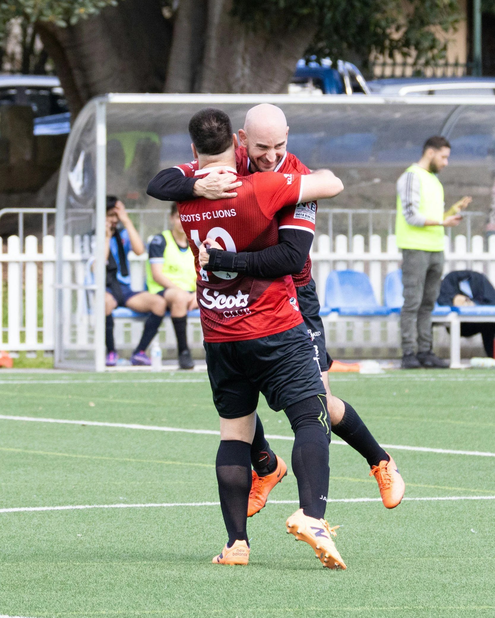 Two soccer players, wearing red jerseys, are hugging on a field, with one smiling. A man in a yellow vest stands in the background, and there are people sitting on a bench behind a white fence.