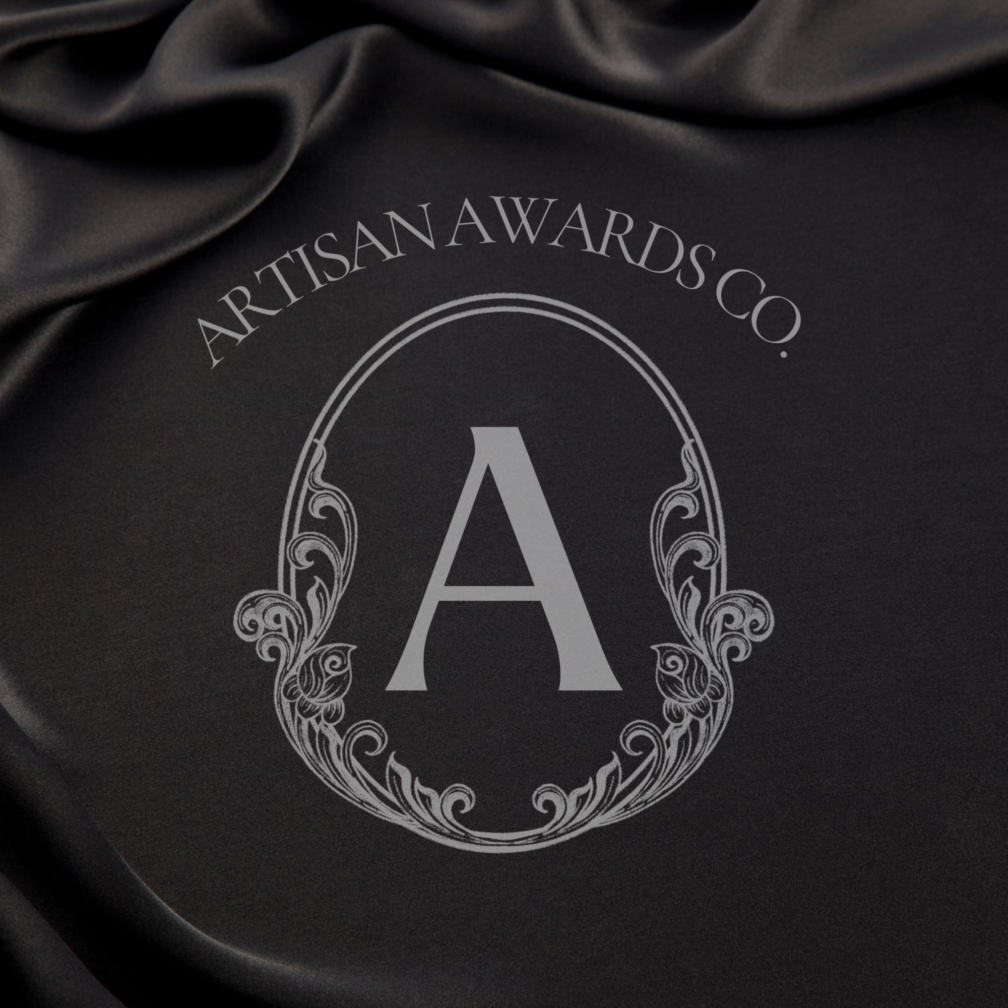 Close-up of a black fabric with a white logo that reads 'ARTISAN AWARDS CO.' in a curved layout, with decorative flourishes around a central large letter 'A'.