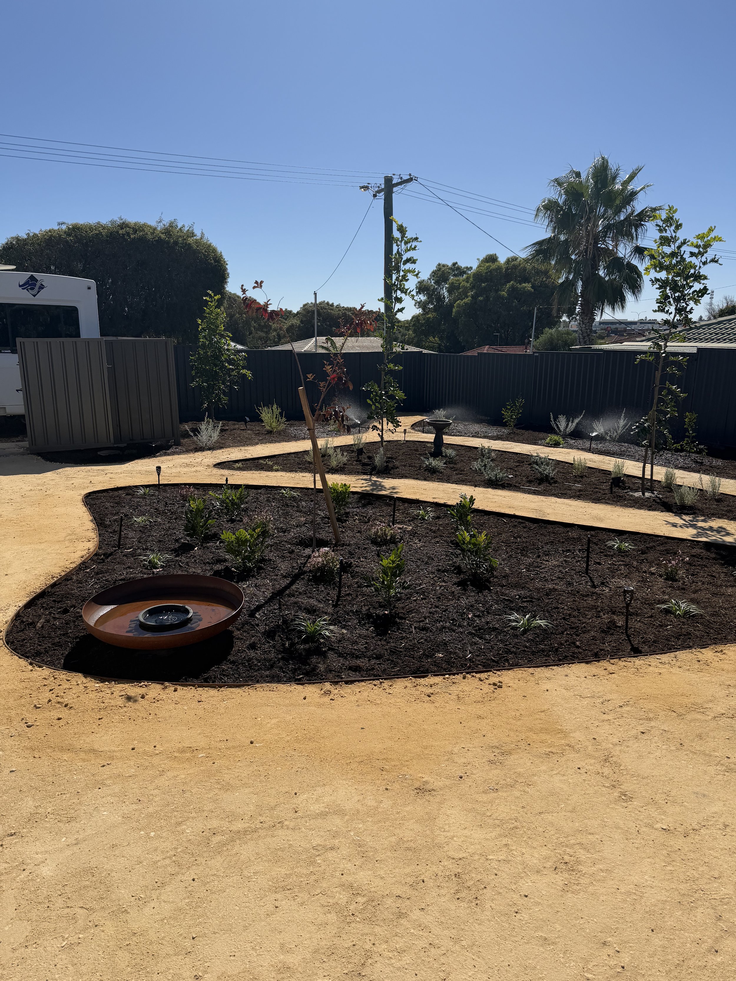 A landscaped garden with small plants and a bird bath, surrounded by a sandy pathway, with trees, a fence, and utility poles under a clear blue sky.