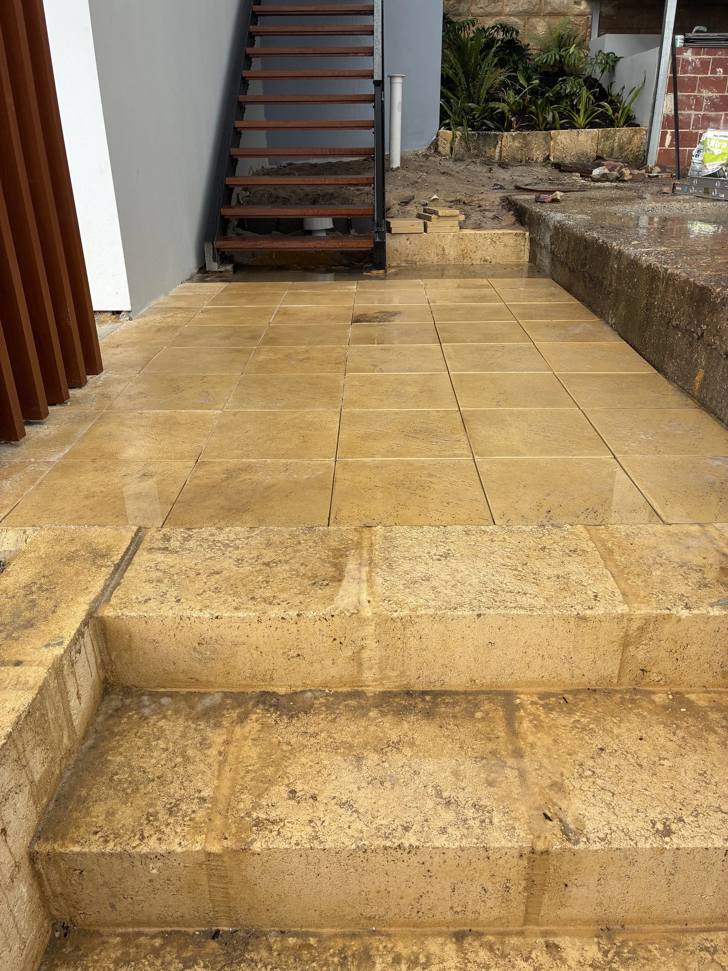 Photo of a construction site showing a partially installed tiled staircase with beige tiles and step edges, leading to an unfinished area with garden plants and construction tools.