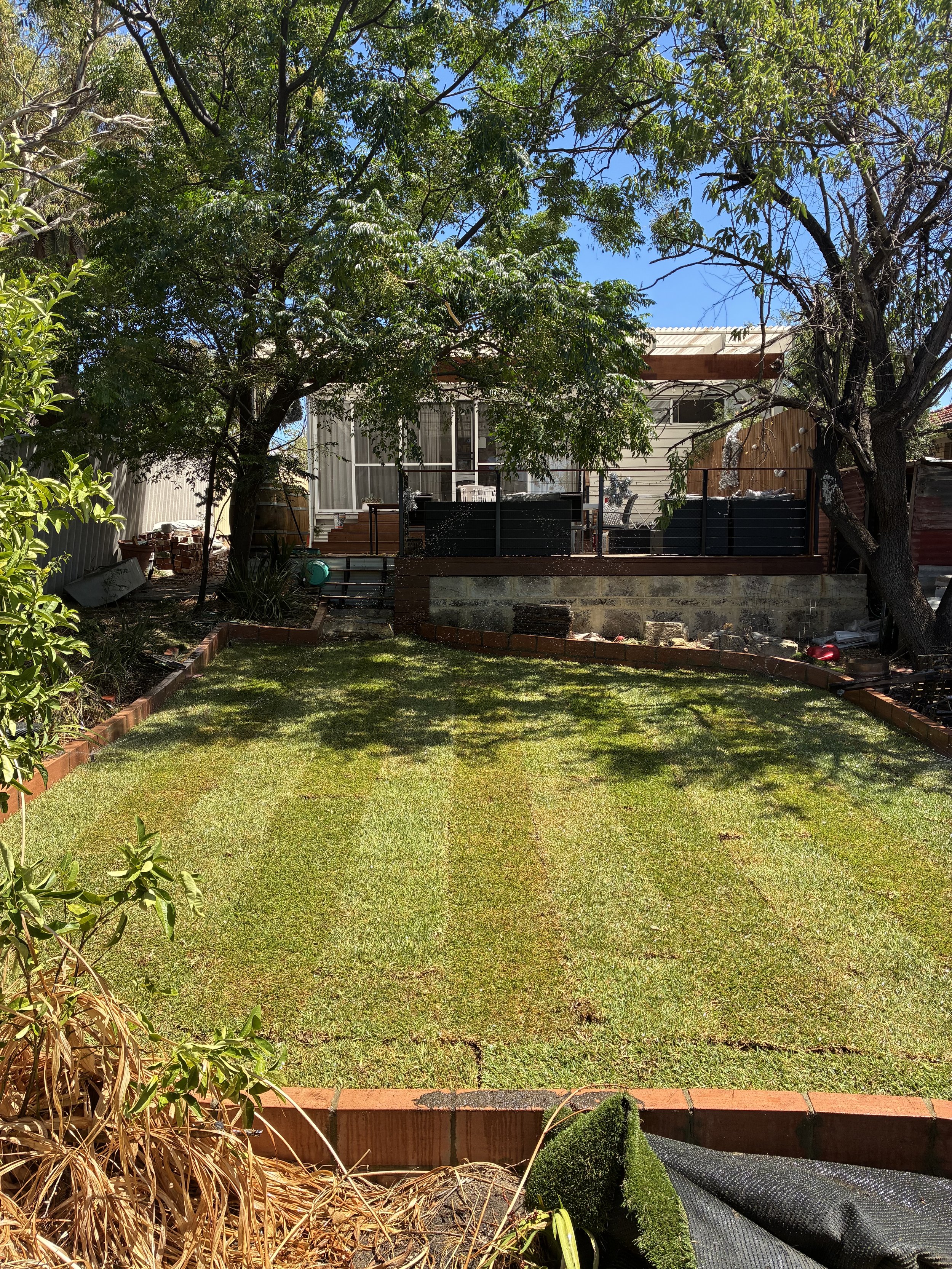 Backyard with a green lawn, trees, a deck with outdoor furniture, and various gardening supplies.