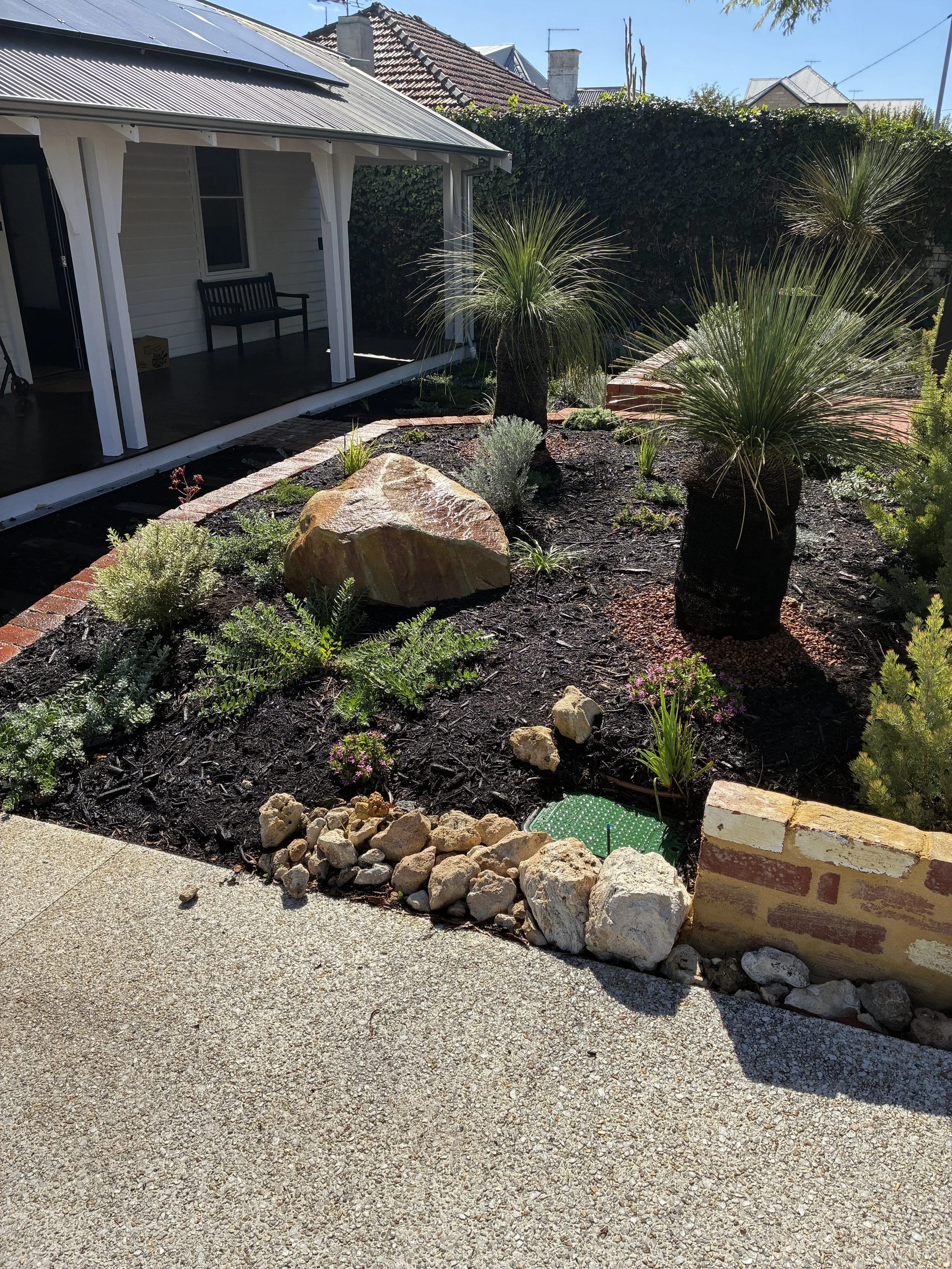 A backyard garden with desert plants, a large rock, and a retaining wall with a brick border. The house with a porch and bench is in the background, along with neighboring rooftops and a clear blue sky.