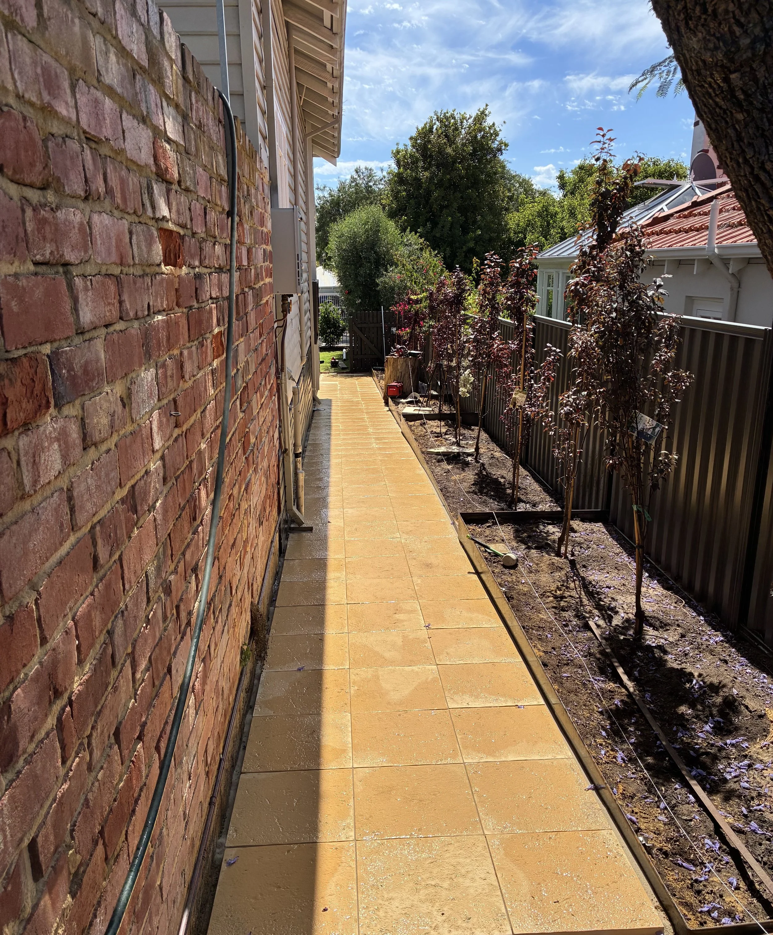 A backyard patio with a brick wall on the left and a row of small trees along the right fence. The patio has a yellowish tile floor that appears wet. The sky is partly cloudy with some sunlight.
