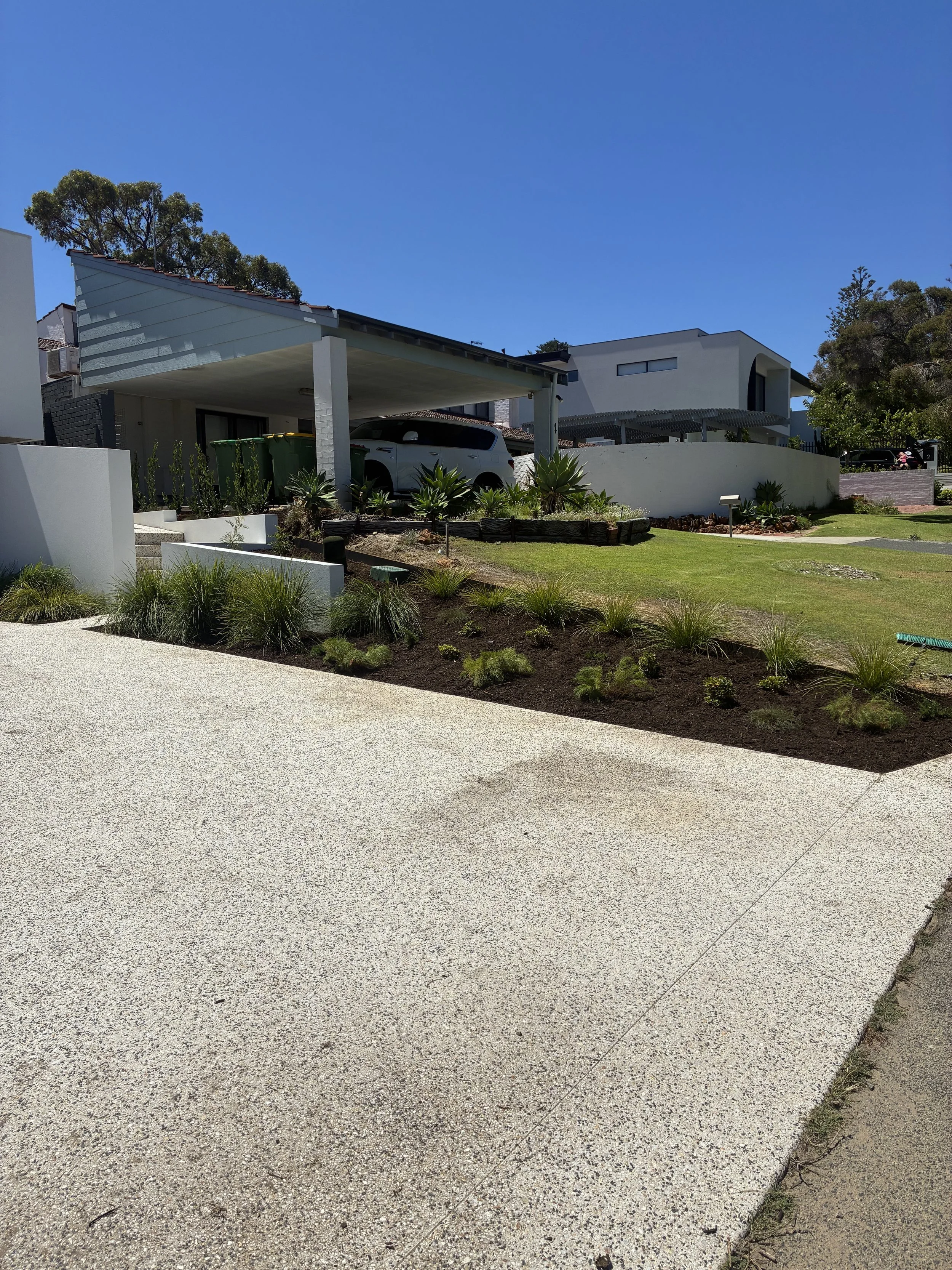 A modern residential house with a carport, a white SUV parked under the carport, and a landscaped front yard with various green plants and a lawn, set against a clear blue sky.