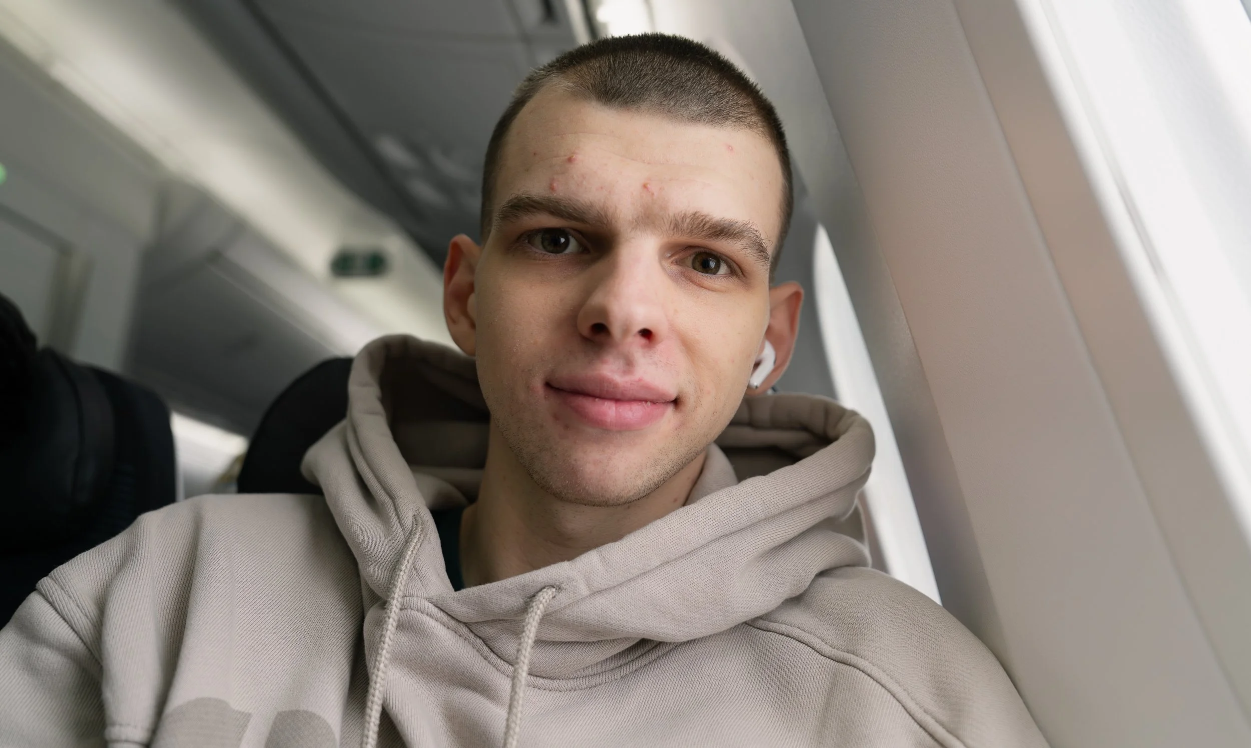 Close-up of a young man with short hair and light skin, sitting next to an airplane window, wearing a beige hoodie and a backpack.