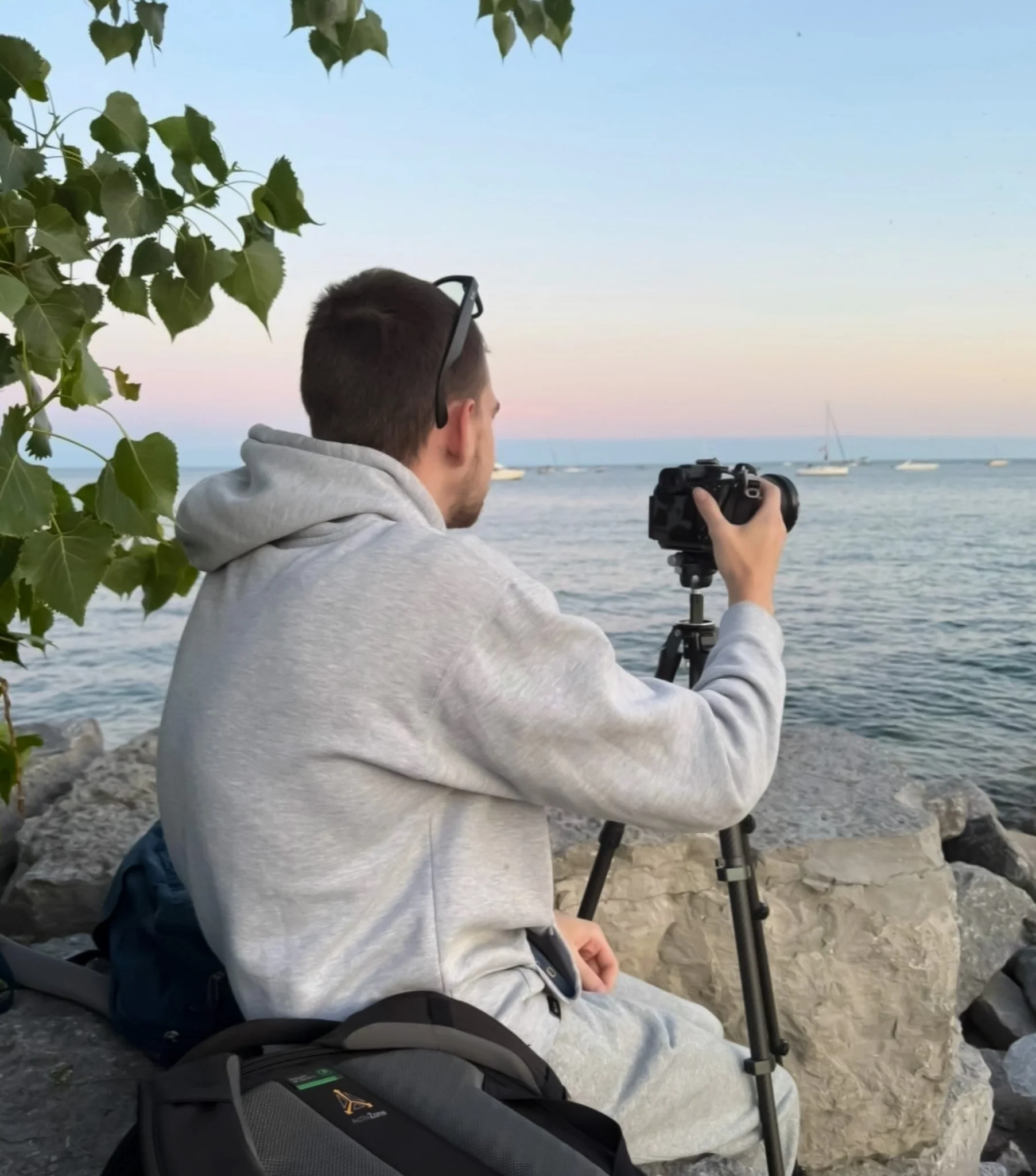 A man sitting on the rocks near the sea, holding a camera on a tripod, with sailboats on the water and a pastel-colored sky in the background.