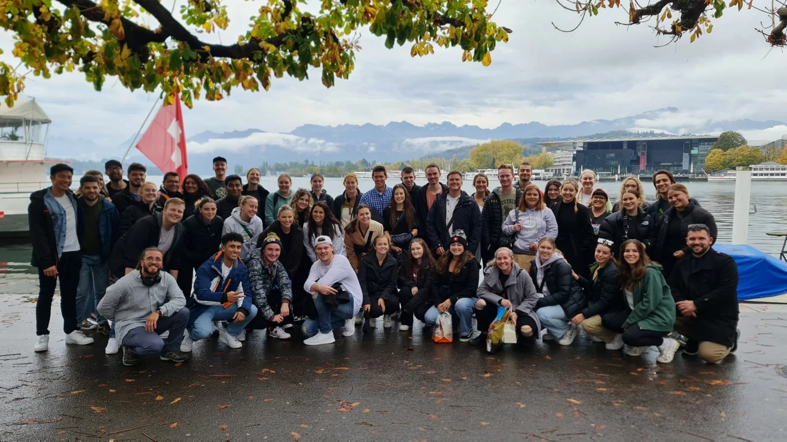 A large group of diverse young people posing outdoors near a body of water on a cloudy day, with mountains and modern buildings in the background.