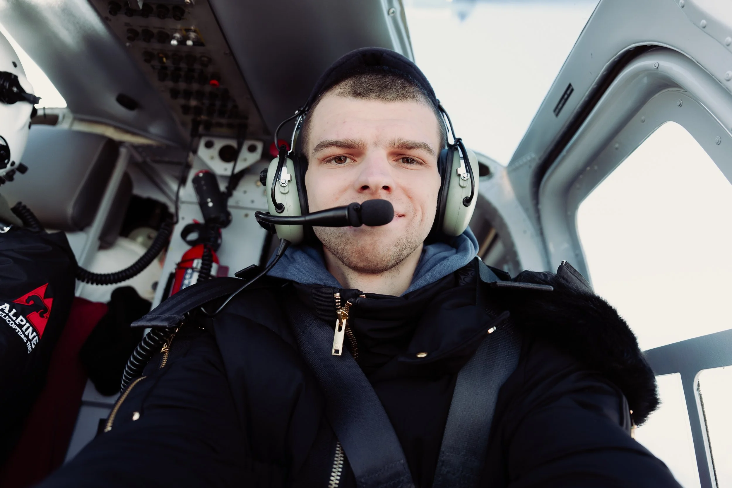 A young male pilot with short dark hair wearing a headset and a black jacket, sitting inside an aircraft cockpit.