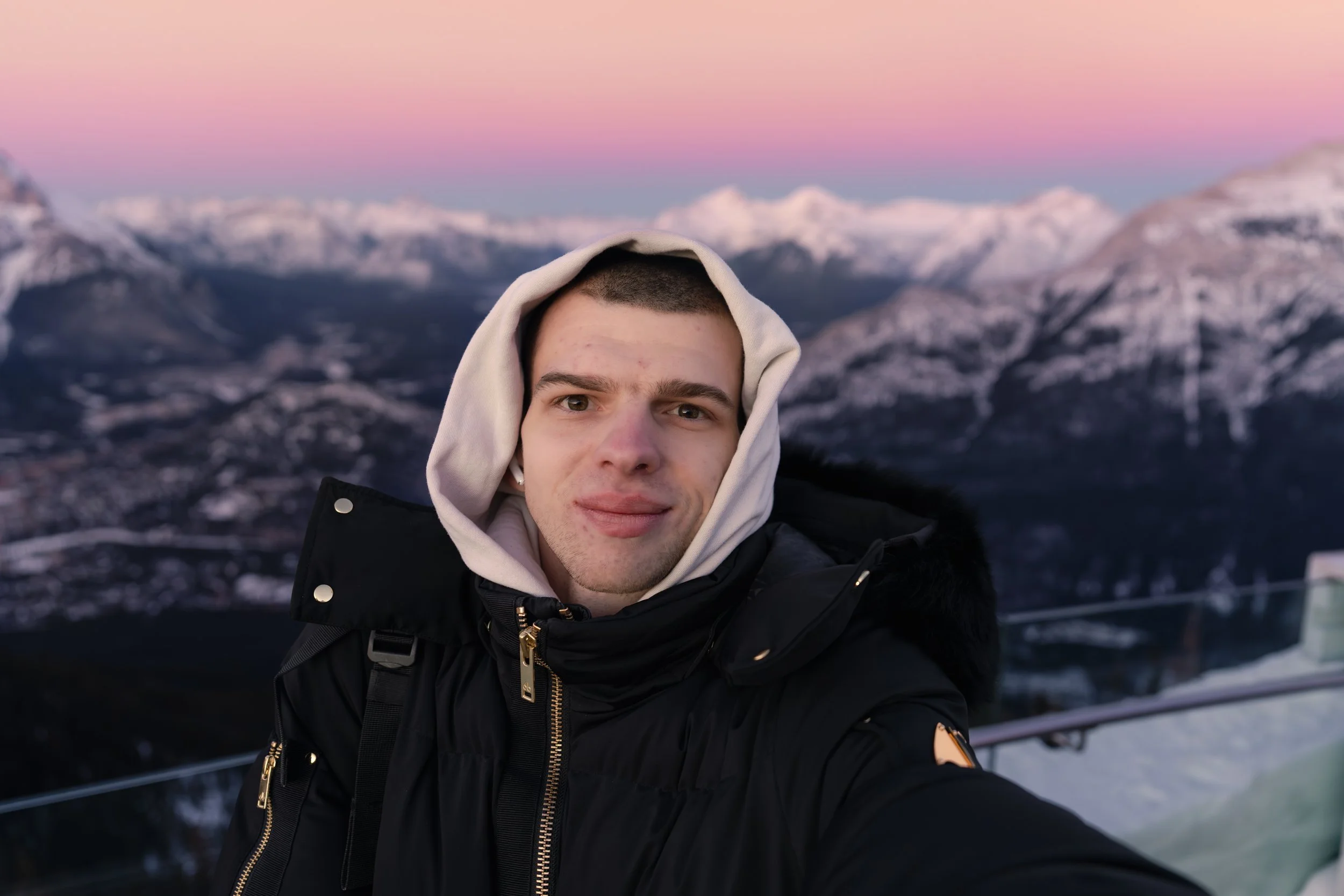 A young man taking a selfie outdoors with snow-covered mountains and a pink sunset sky in the background. He is wearing a black jacket with a hoodie and has a slight smile.