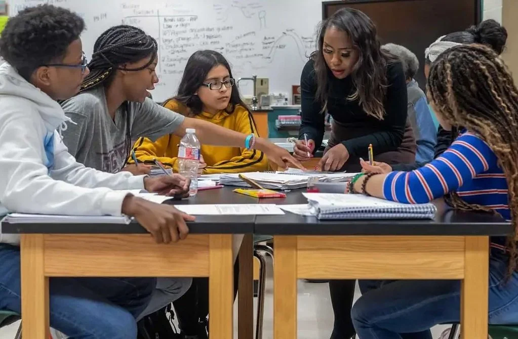 A diverse group of students gathered around a table in a classroom, engaged in a discussion with a female teacher, who is teaching them how to be successful.