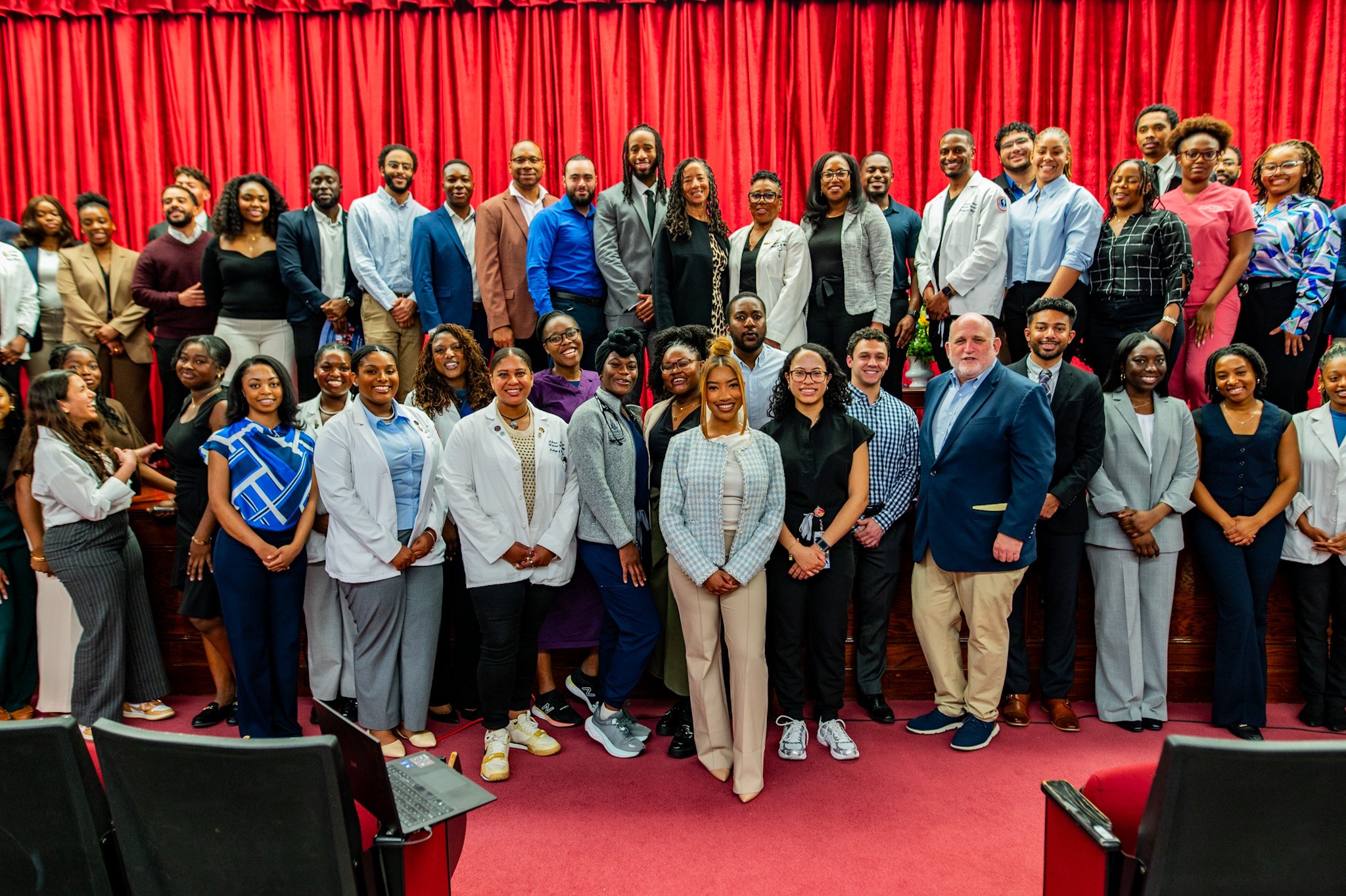HUMAA scholarship team and recepients standing together on stage in front of red curtain, smiling for a group photo.