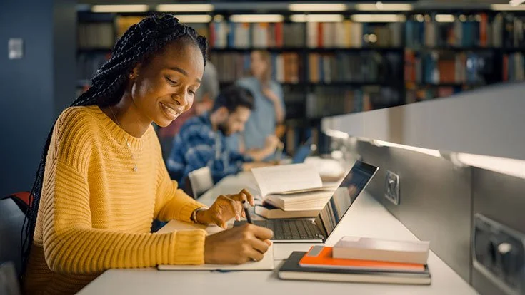 Caribbean girl writing on a notepad in front of laptop working on her financial future and success.