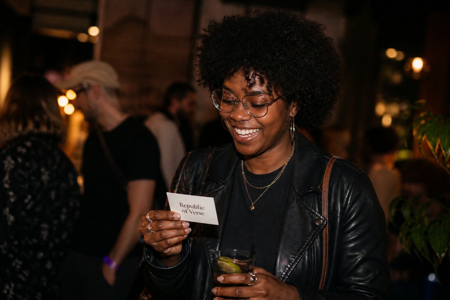 A smiling woman with glasses and a septum piercing holding a drink and a card that reads 'Republique of Verse' at a social gathering or event.