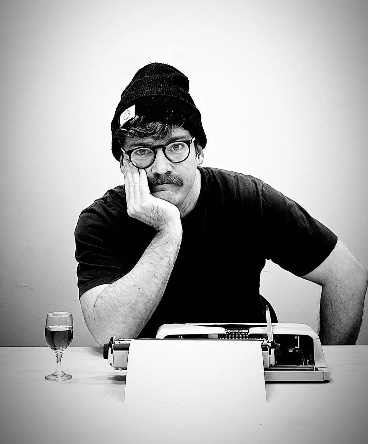 Black and white photo of a man with glasses and a mustache, wearing a beanie, sitting at a table with a typewriter, a glass of water, and a blank piece of paper, resting his face on his left hand with an expression of boredom or contemplation.