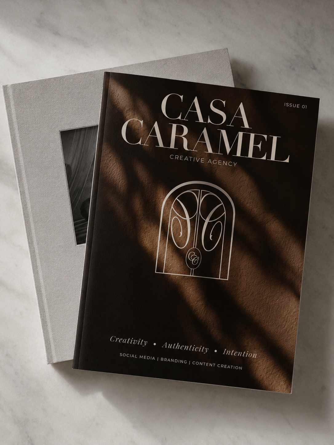 A magazine titled CASA CARAMEL on top of a white book with a photo visible through a cutout. The CASA CARAMEL magazine cover is dark brown with white text and a minimalist illustration of a door.