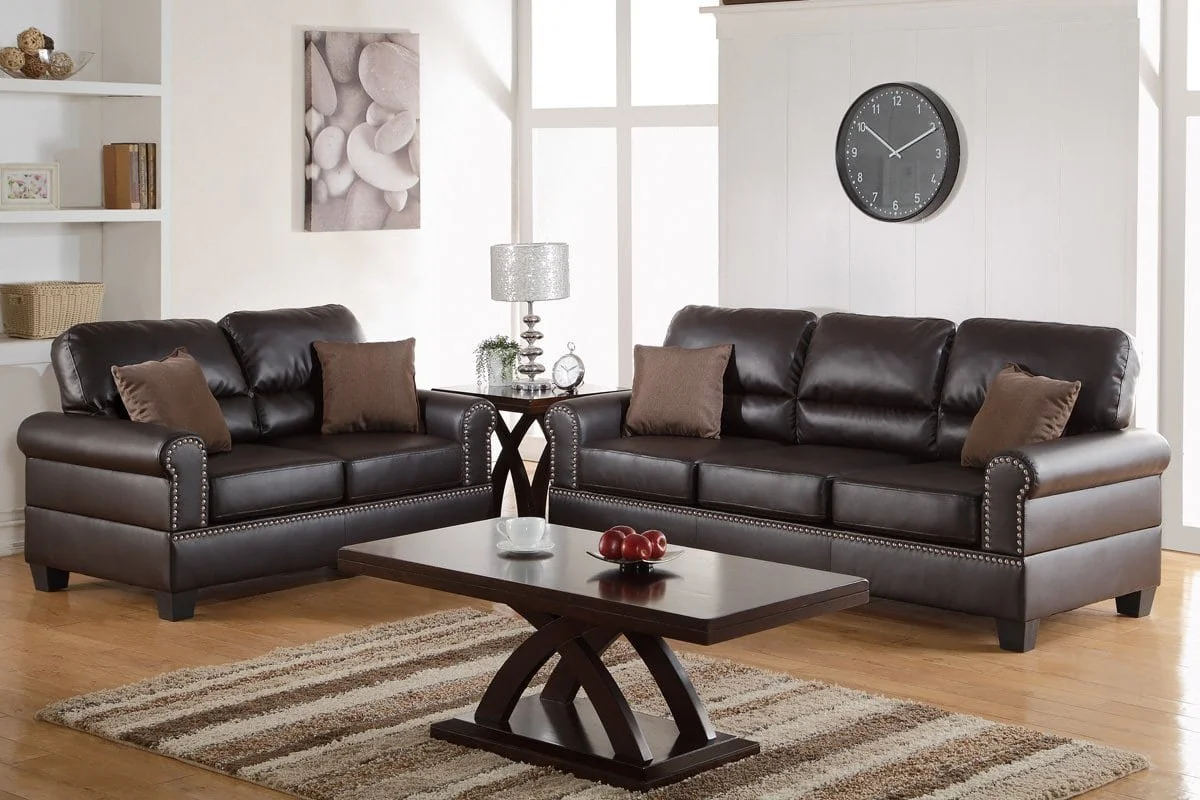 A living room with two dark brown leather sofas, a wooden coffee table with a bowl of apples, a side table with a lamp, and a wall clock. The room has light-colored walls, large windows, and a striped area rug on the hardwood floor.