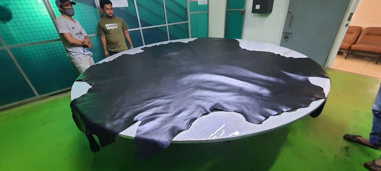 People standing around a large, oval table covered with a black leather hide in a room with green floor and blue-green partition walls.