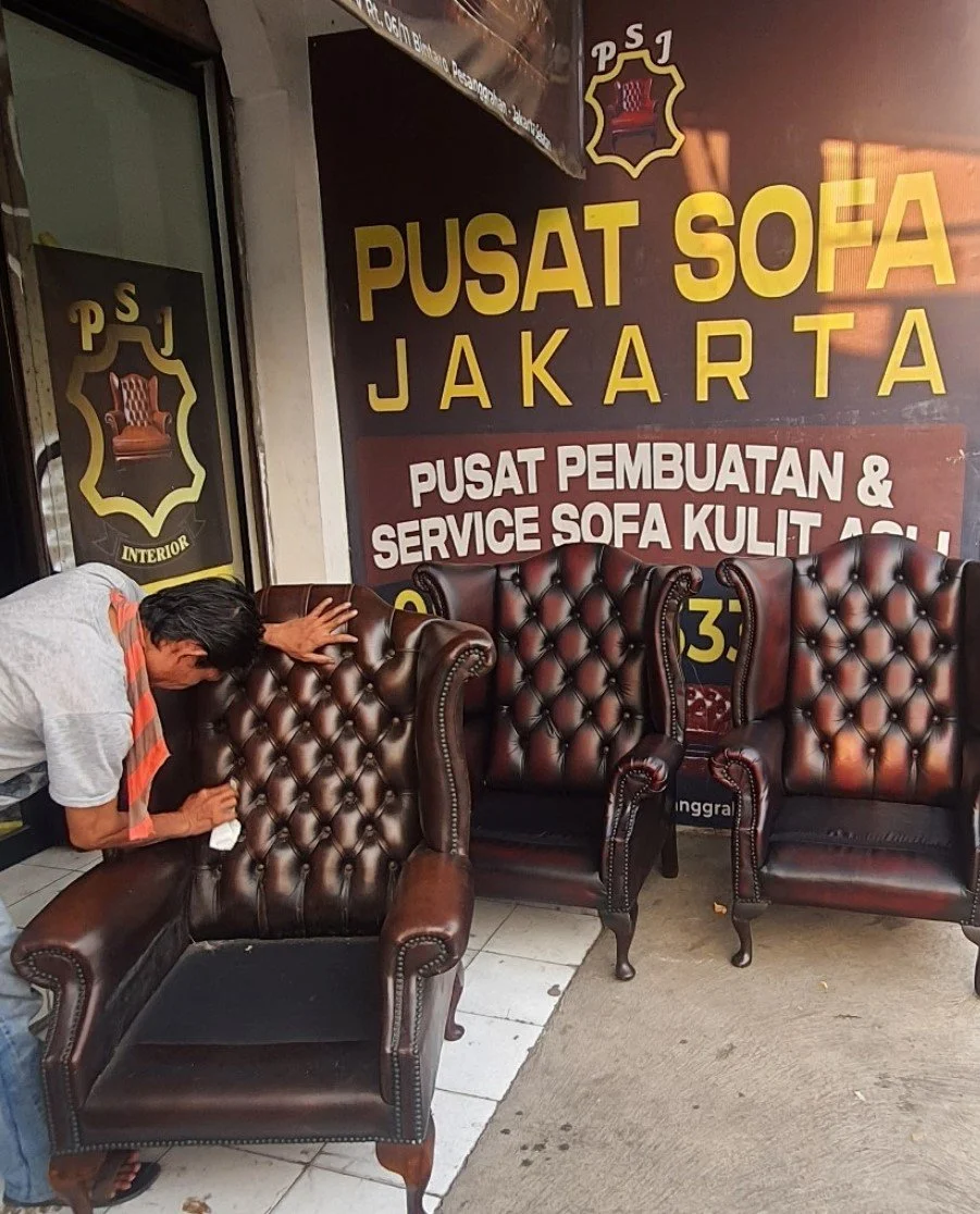A man cleaning a large brown leather tufted sofa outside a furniture shop with a sign that reads Pusat Sofa Jakarta, which is a sofa manufacturing and service center in Jakarta.