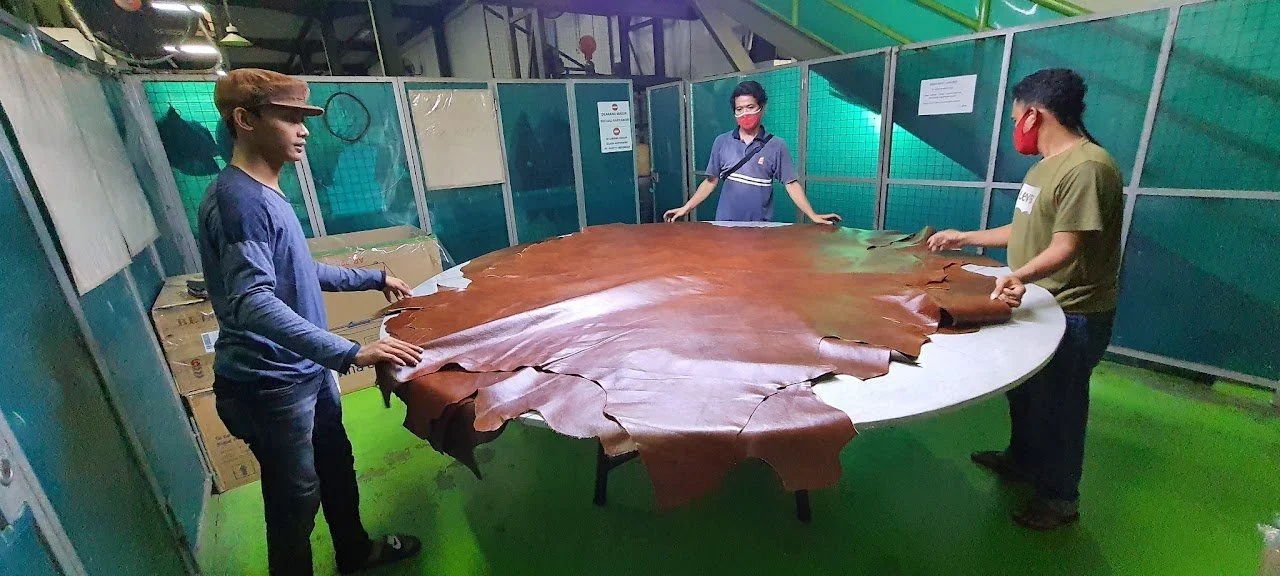 Three workers in a factory, all wearing masks, handling large pieces of brown leather on a table.