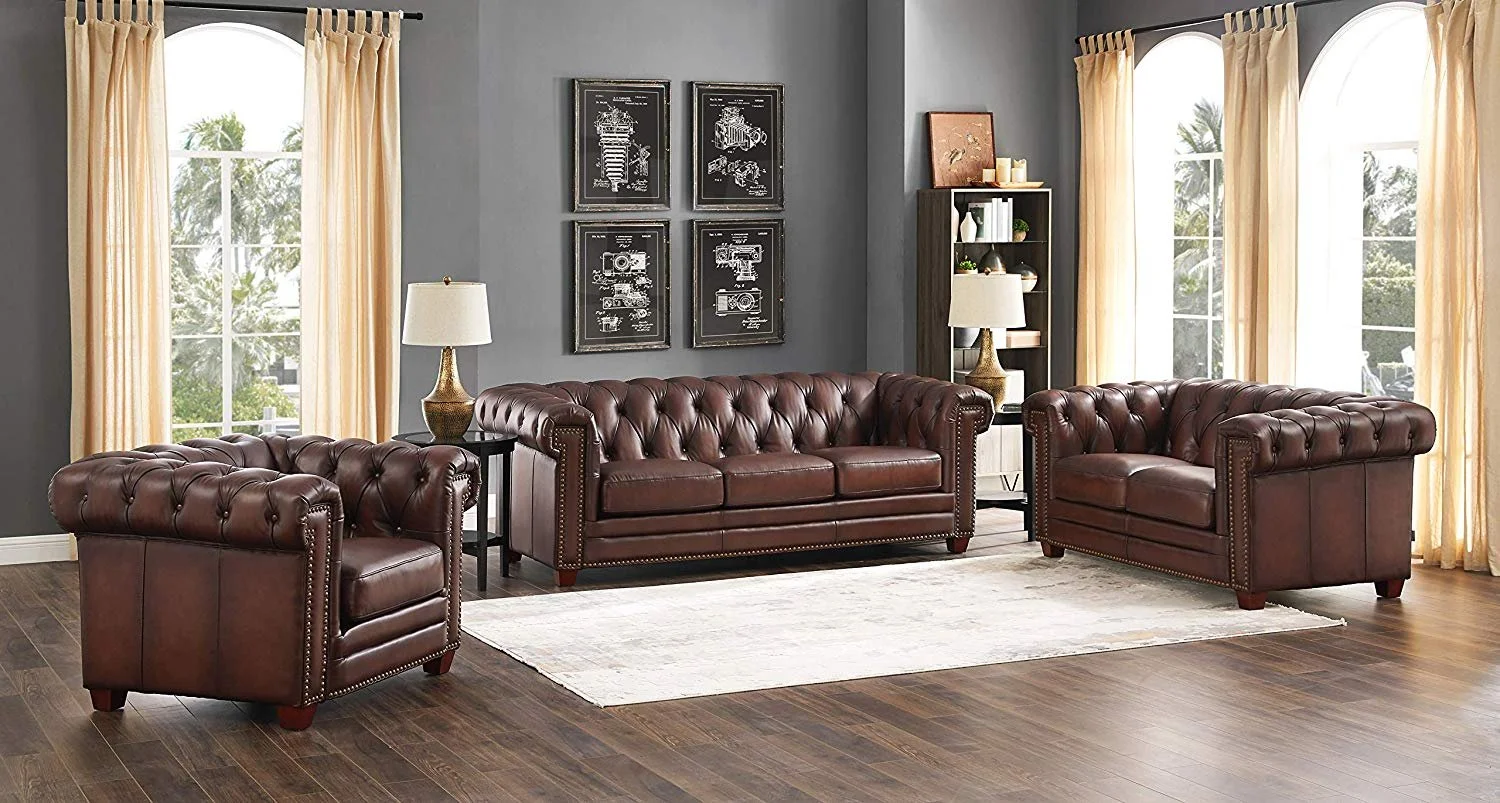 Living room with gray walls, three large windows with beige curtains, dark hardwood floors, a cream area rug, and a set of three brown leather tufted sofas. There are two side tables with lamps and wall art featuring blueprint-style drawings.