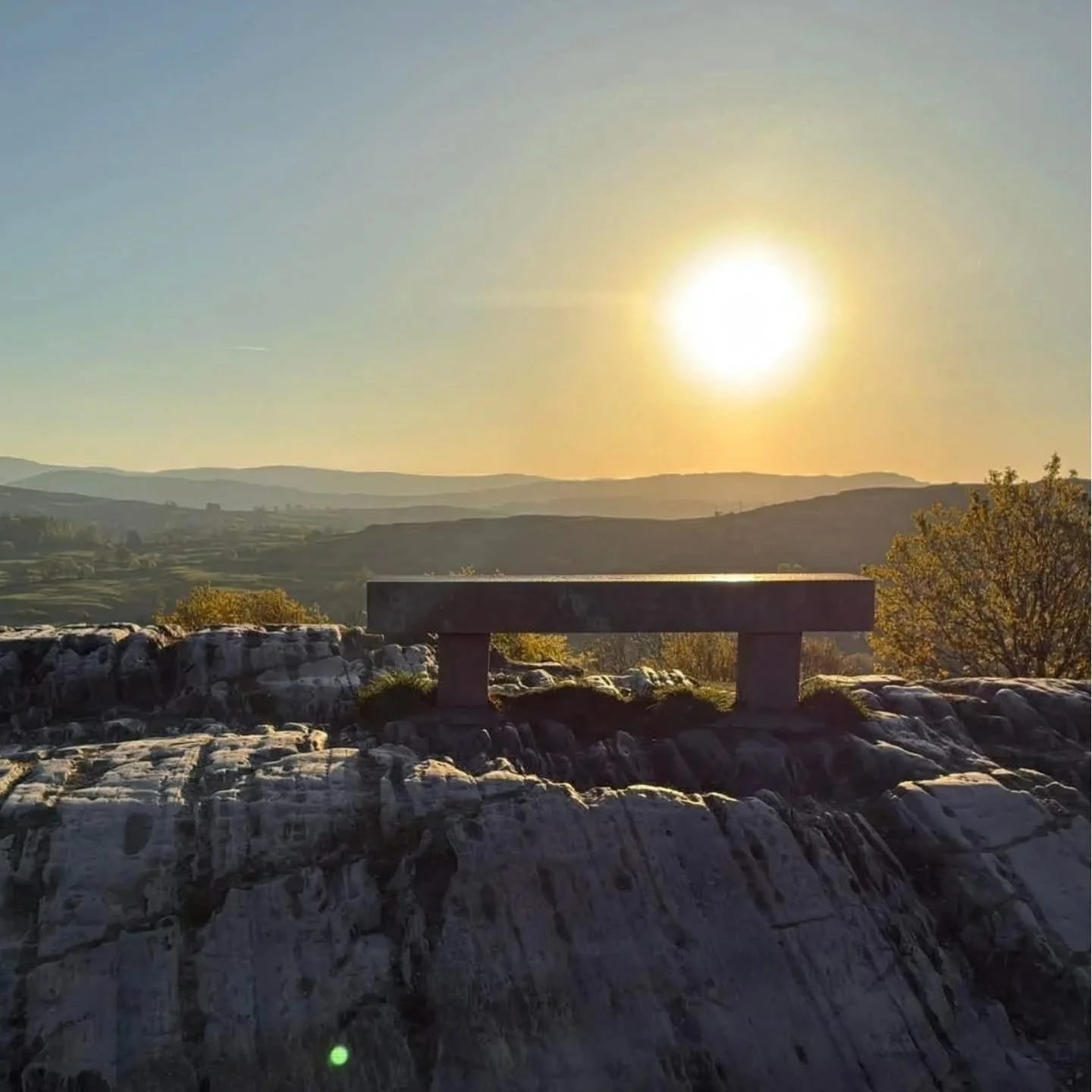 A stone bench on a rocky surface with a scenic view of rolling hills in the background, under a bright sun in the sky.