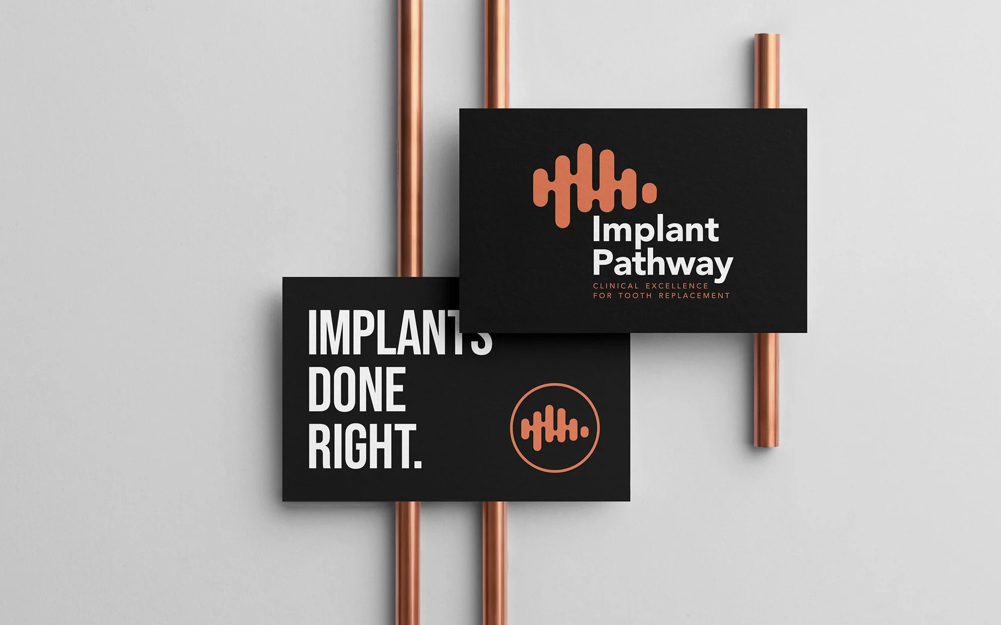 Black sign with orange and white text reading "Implant Pathway" and "Clinical Excellence for Tooth Replacement", with a logo resembling a sound wave. Next to it, part of another sign with the text "Implants Done Right" in white on a black background. The signs are mounted on copper pipes against a light gray wall.