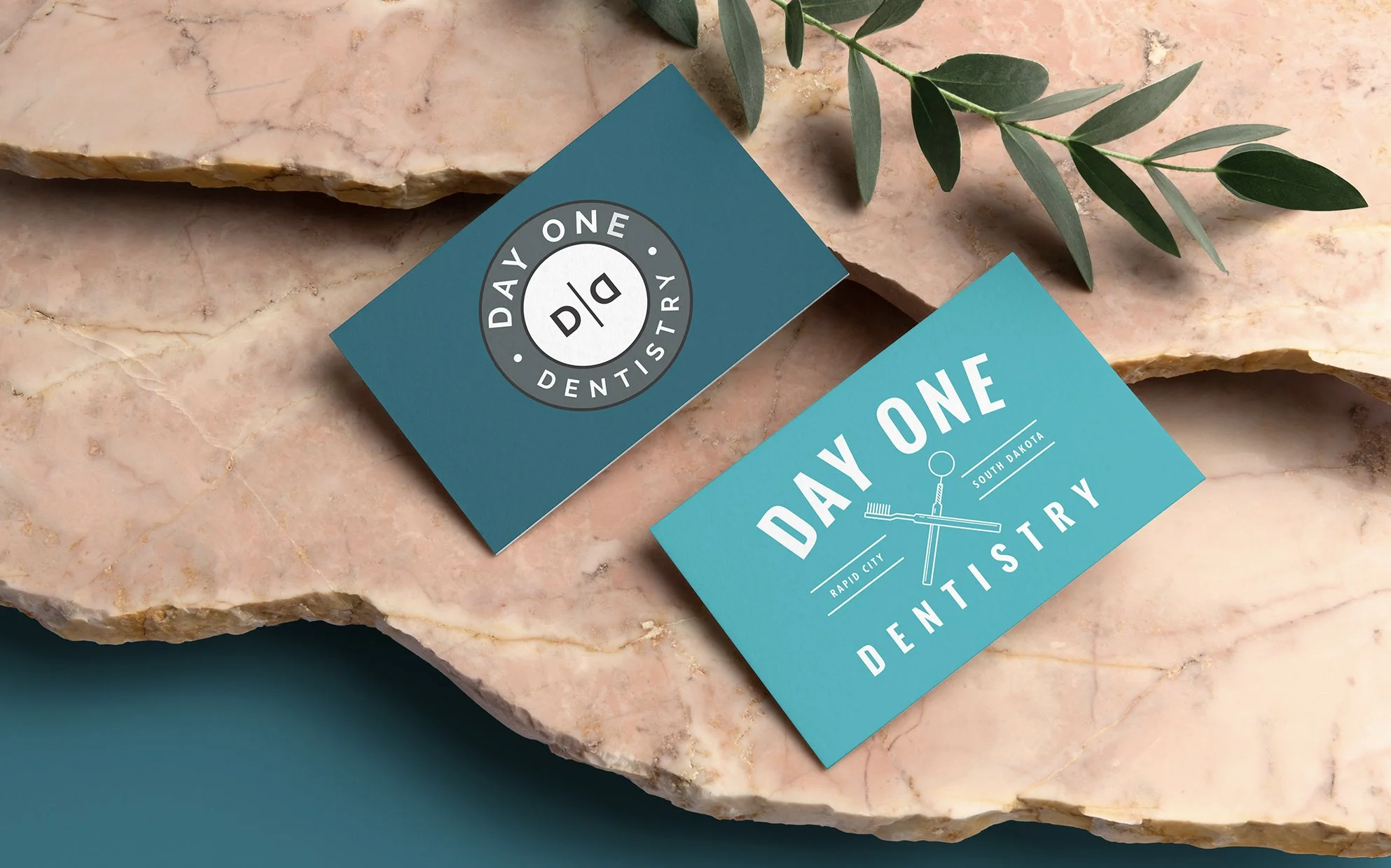 Business cards for Day One Dentistry placed on a large marble slab with a green leafy branch nearby.