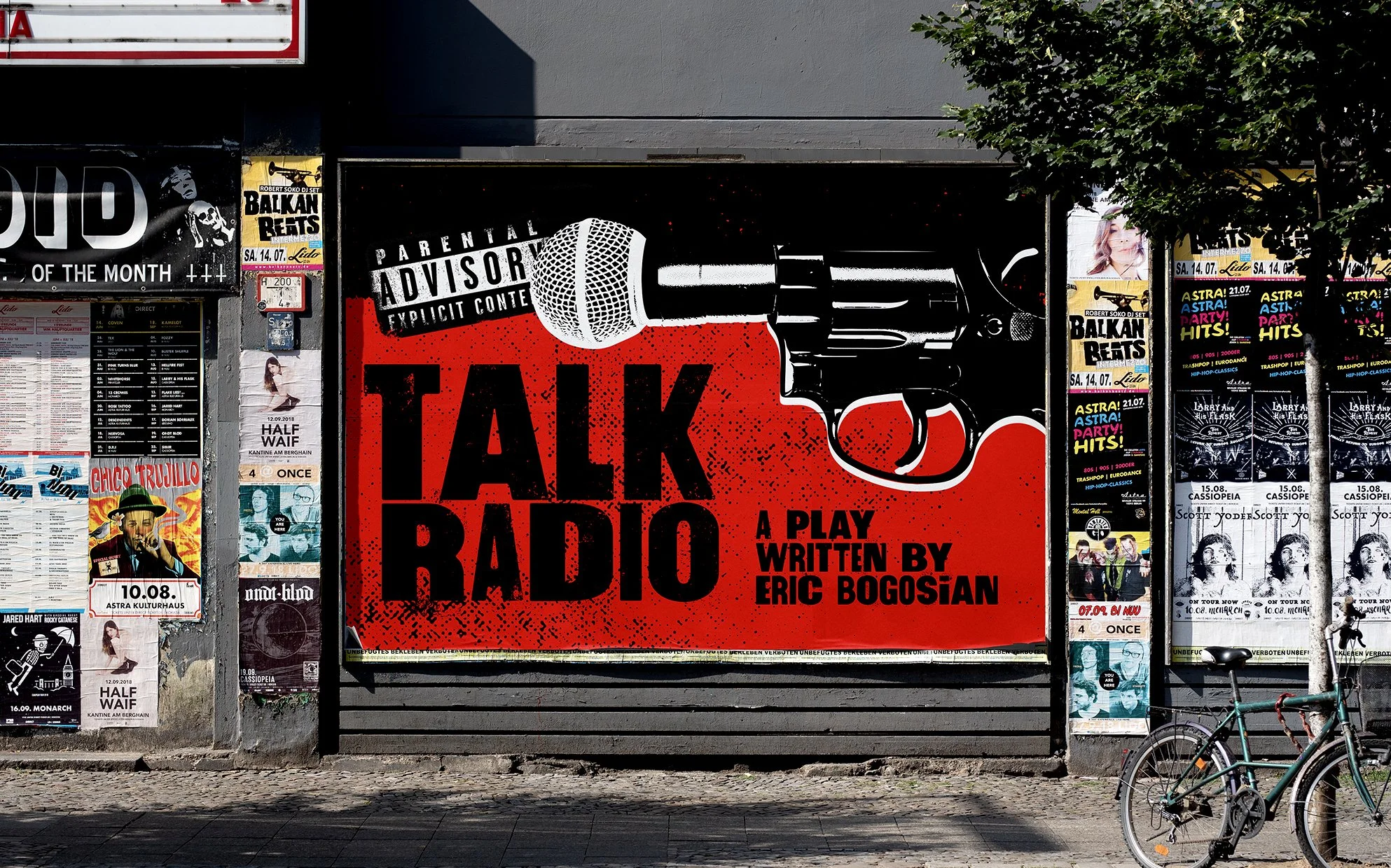 A city street scene with a large red poster advertising a play titled 'Talk Radio,' written by Eric Bogosian, with a black and white microphone illustration. Surrounding posters promote music events, theater performances, and other entertainment.