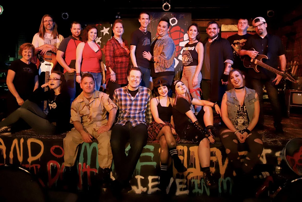 Group of people on a stage, some holding musical instruments, in a dimly lit venue with graffiti-style art in the background.