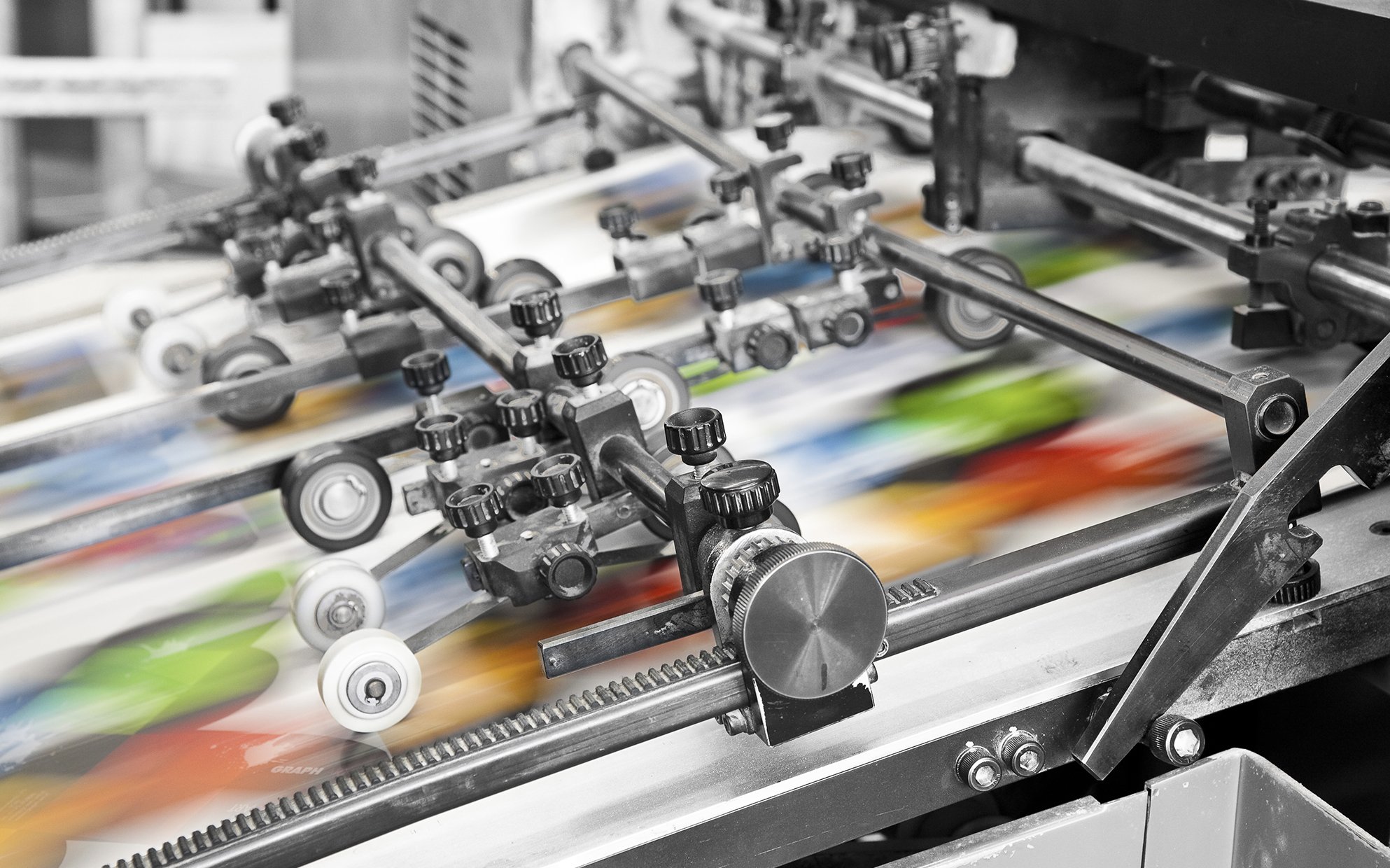 Close-up of a printing press machine in action, printing colorful images on paper with various mechanical parts and rollers.