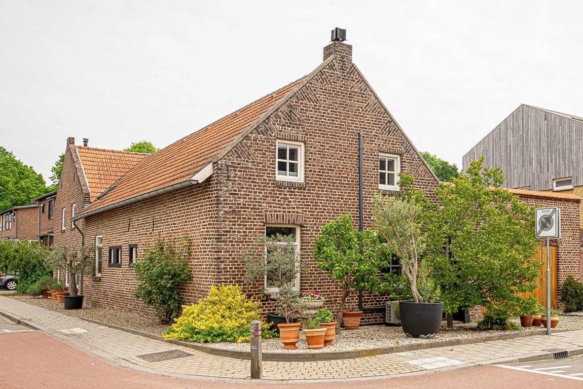 Een traditioneel rood bakstenen huis met een houten dak en meerdere kleine planten en boompjes voor de gevel.