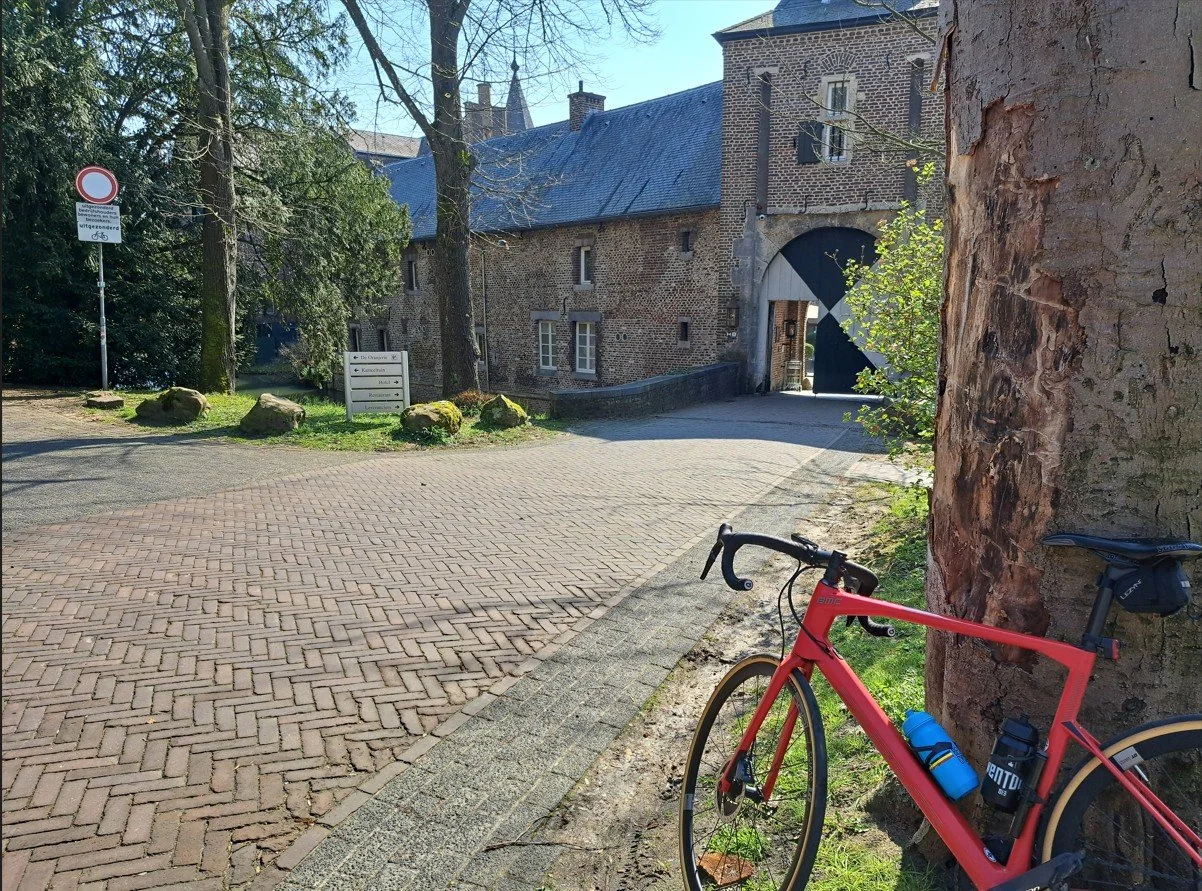 Bromfietsparkeren naast een grote boom voor een historische stenen muur en een boogpoort in een dorp. Een wegwijzer en een verkeersbord zijn zichtbaar.