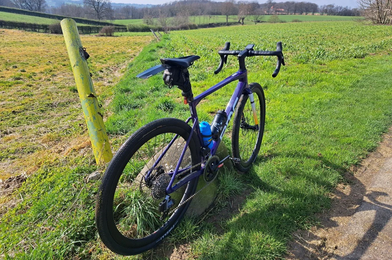 Een racefiets met een paar waterflesjes op een graslijn langs een veld, met een heuvelachtig landschap en bomen op de achtergrond.