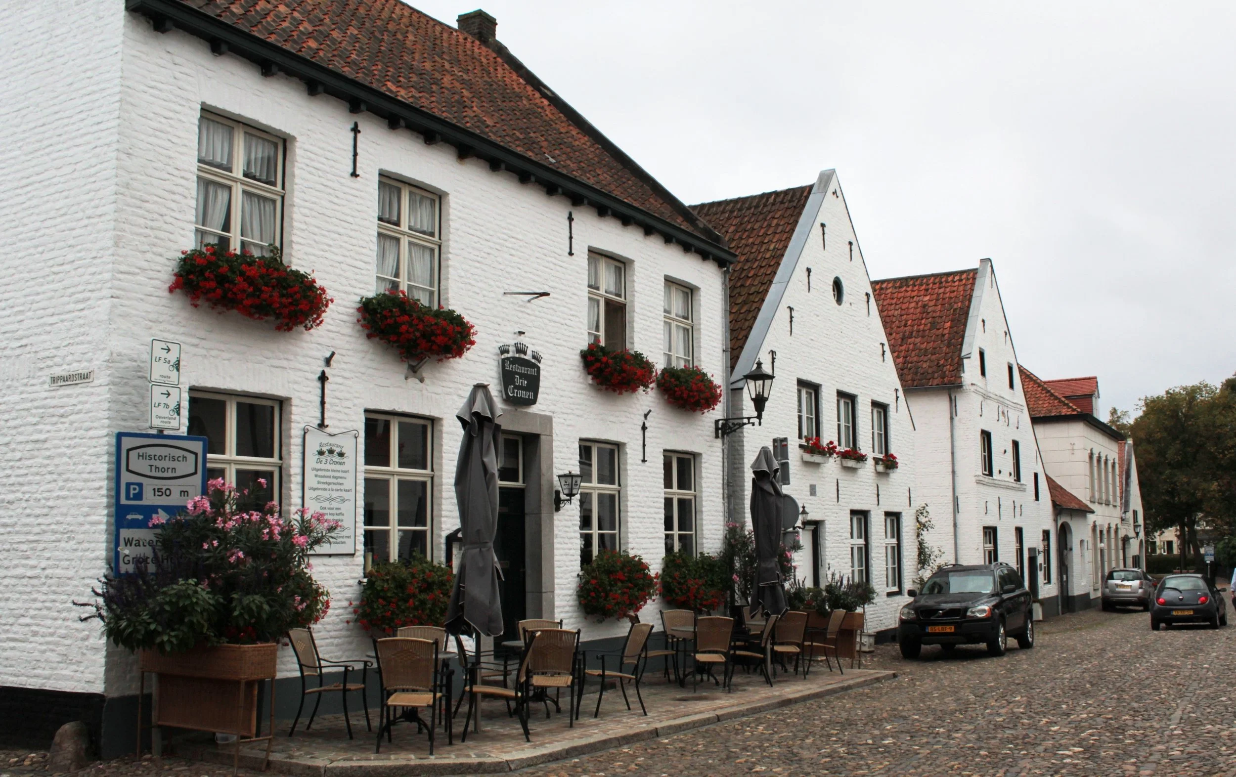 Uitzicht op witte gebouwen met rode daken en ramen met bloempotten vol rode bloemen. Er staan buitenmeubels en paraplu's op de straat, met enkele geparkeerde auto's op de straat. Borden en straatnaamborden zijn zichtbaar.
