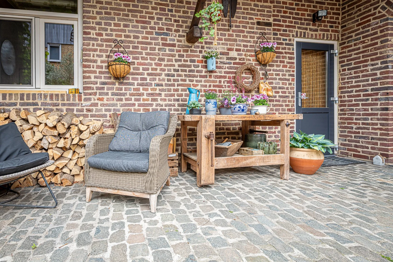 Een buitenruimte met een bakstenen muur, een raam en een deur. Er ligt een stapel houtblokken, er zijn twee stoelen en een houten tafel met planten en decoraties. Daarnaast staan planten in potten en hangen bloemversieringen aan de muur.