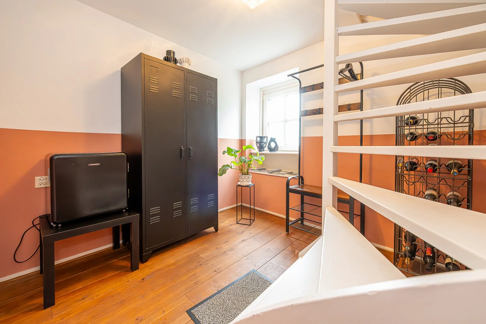 Een kamer met houten vloer, een zwarte kast, een wijnrek met wijnflessen, een plant op een kleine tafel, een raam en enkele decoratieve objecten.