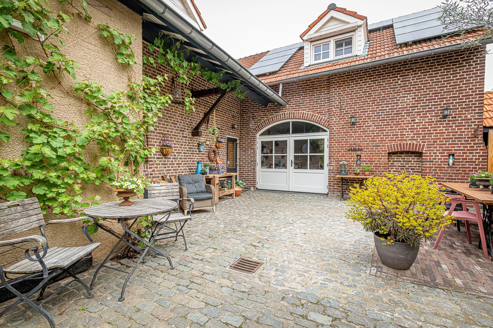 Een gezellige tuin patio met een keramische tegelvloer, een rotan bank met kussens, een houten tafel, en bloempotten met bloemen, omringd door een bakstenen muur en een huis met zonnepanelen op het dak.