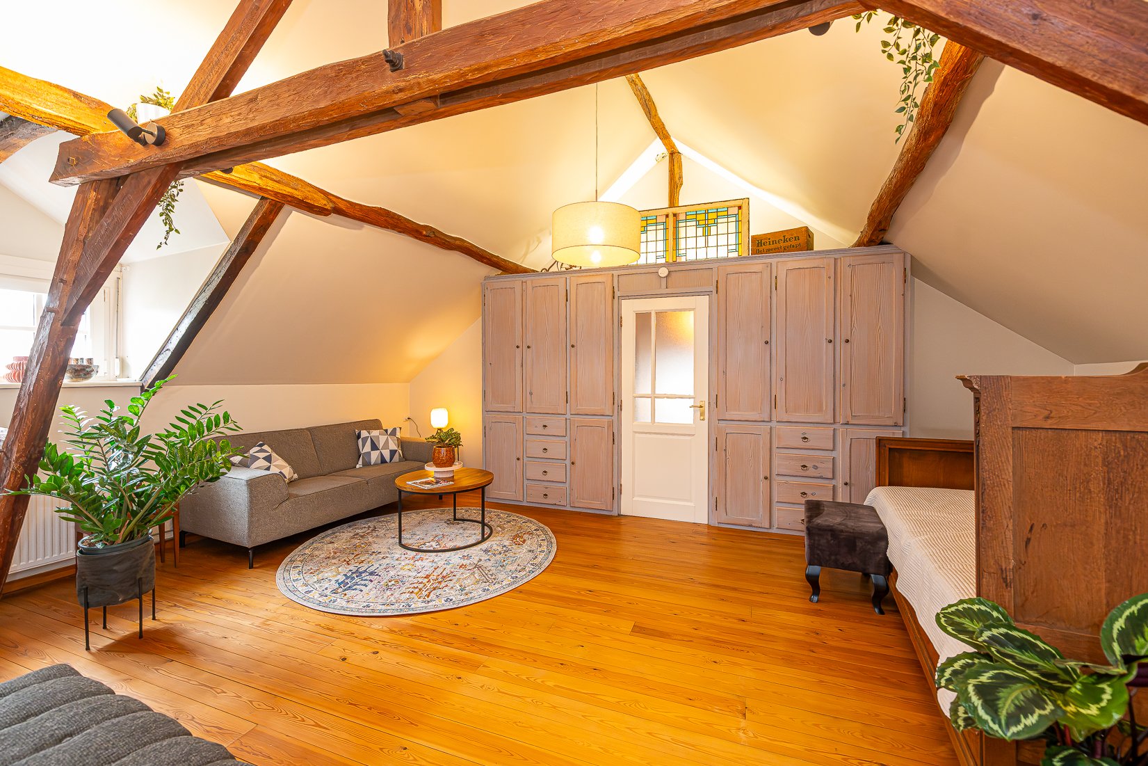 Een woonkamer met houten vloeren en balken, een grijze bank met vierkanten kussens, een ronde salontafel met een tas en tijdschrift, een kast met lades en deuren, een deur met glas, een plantenpot met groene bladeren, een ronde vloerkleed, en een gro