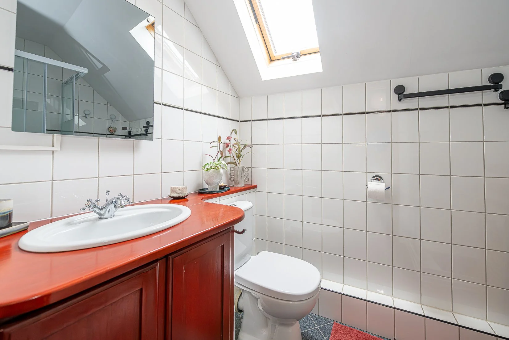Een kleine badkamer met witte tegels, een dakraam, een spiegel, een wasbak met een houten kast, een toilet, en enkele planten en decoraties.
