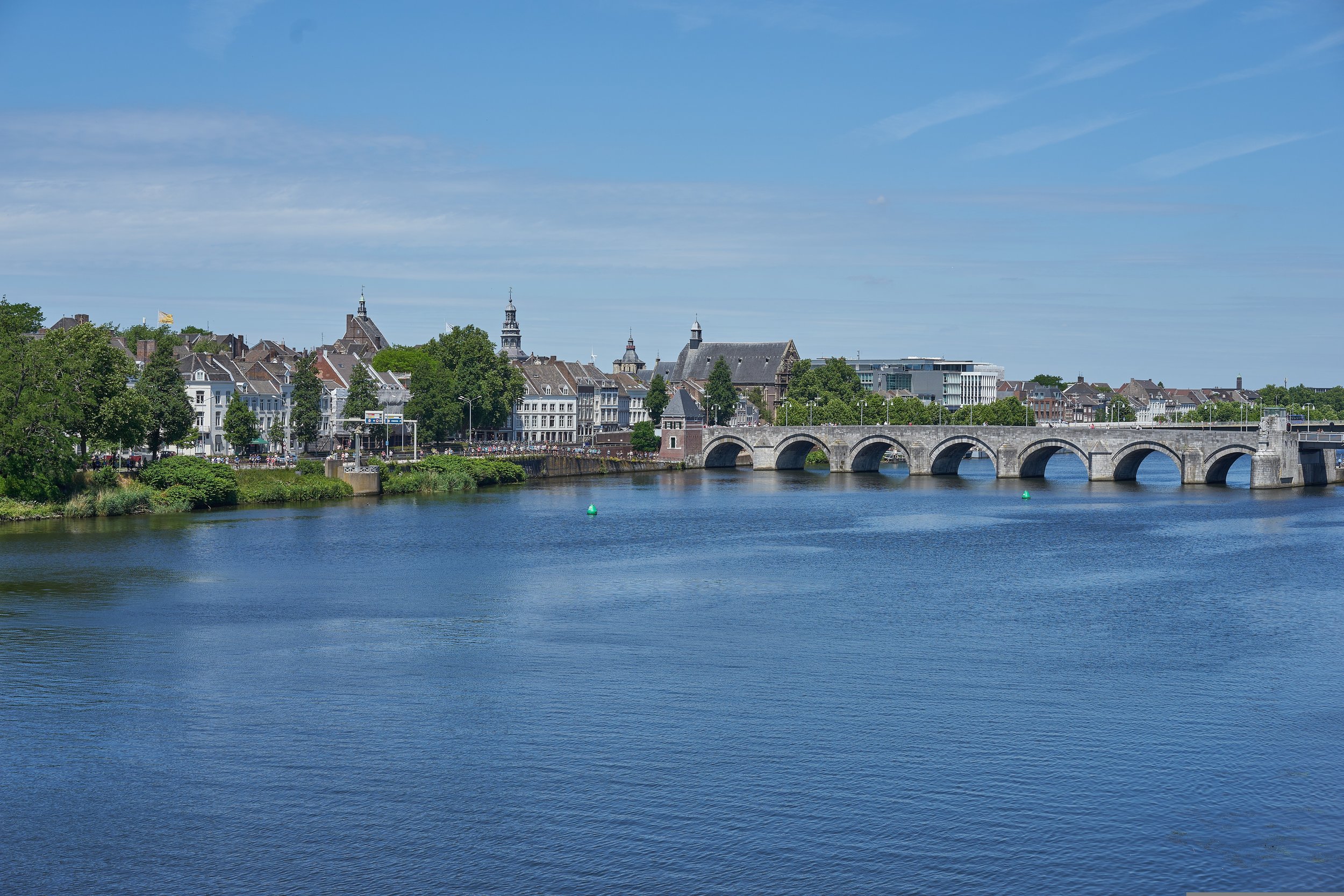 Een stadszicht met een oude stenen brug over een rivier en historische gebouwen en kerktorens op de achtergrond, onder een blauwe lucht.