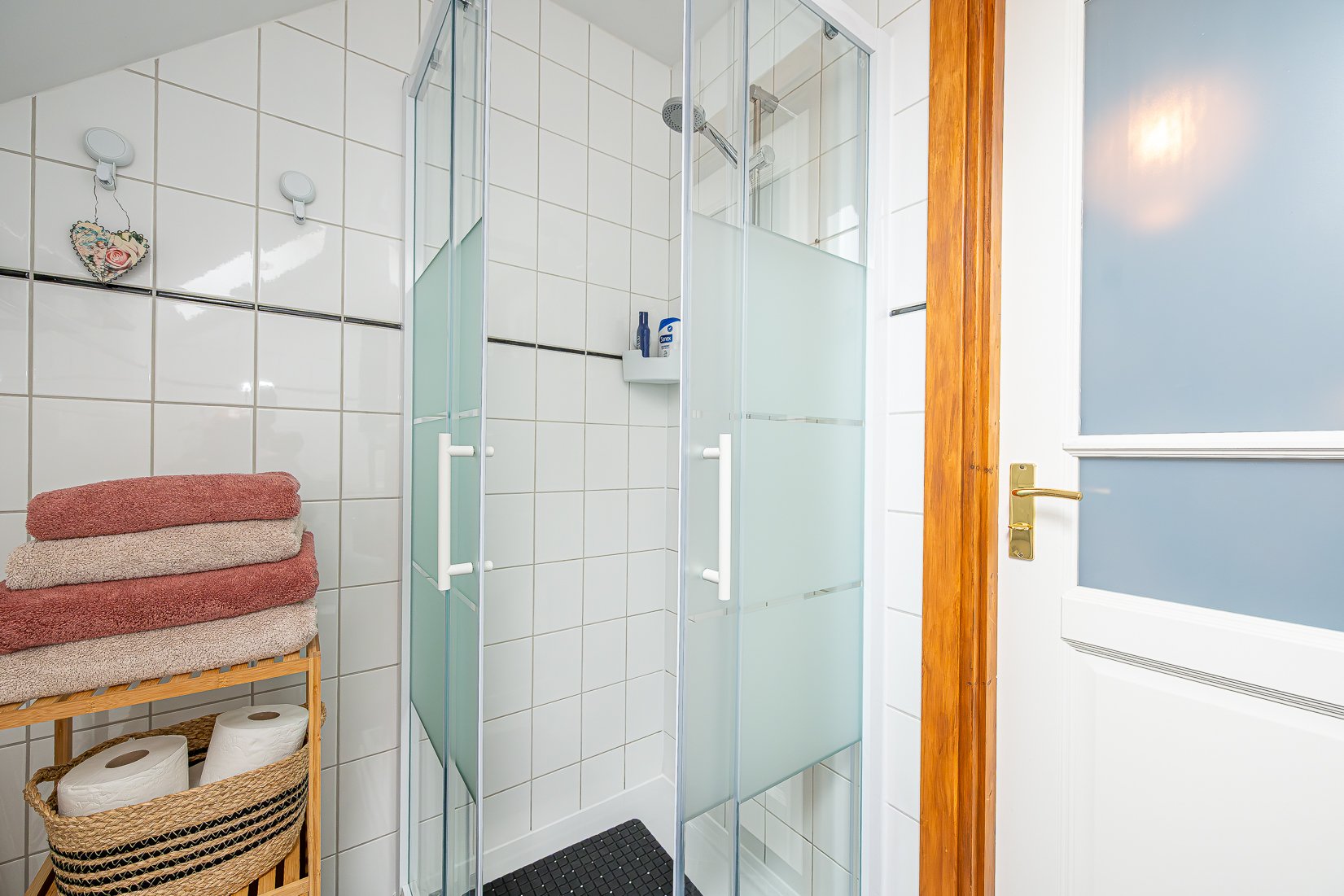Een badkamer met een nette inloopdouche, witte tegels op de muur, een houten rek met handdoeken en toiletrolletjes, en een sierlijke deur met een gouden klink.