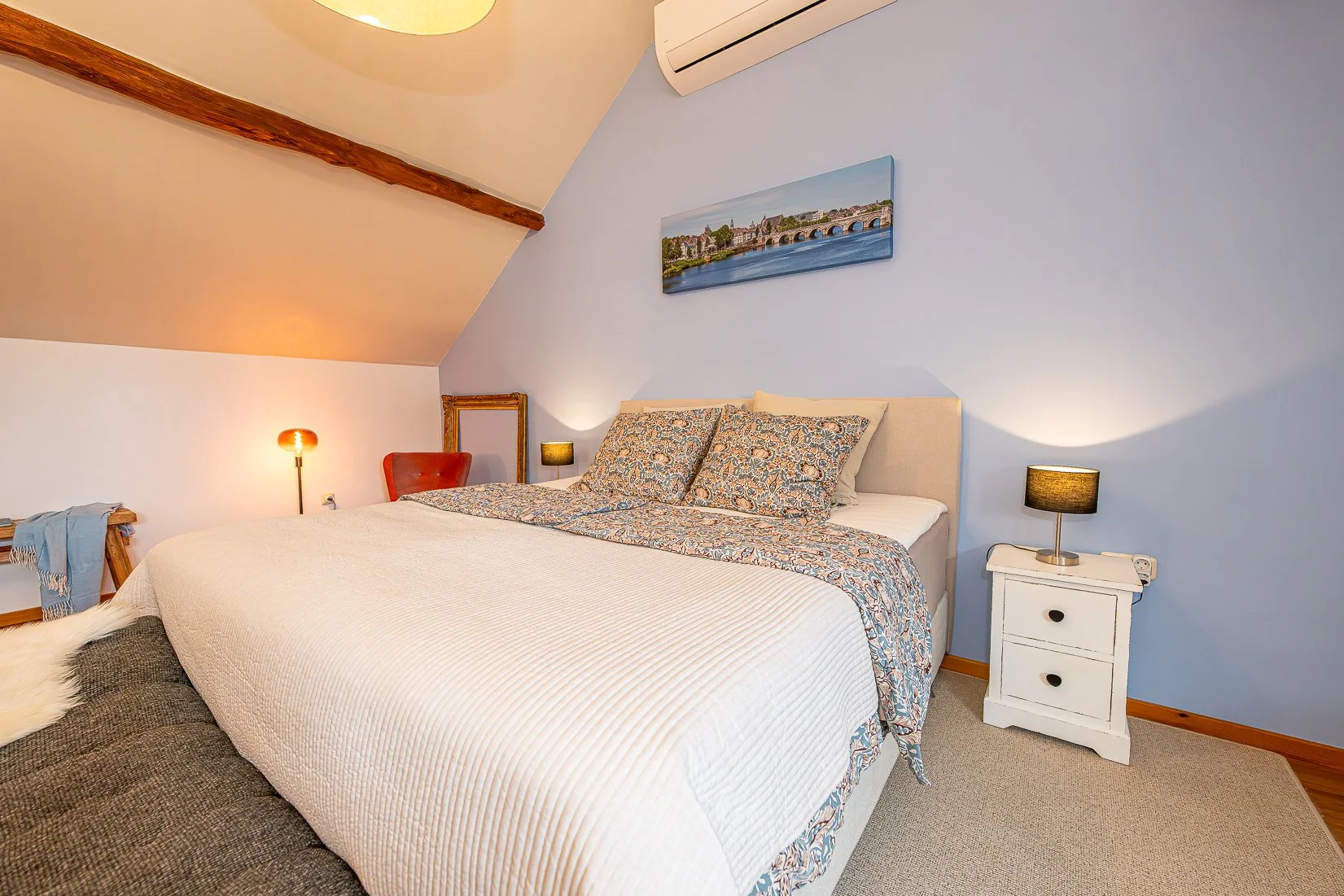 Slaapkamer met wit bed, blauwe muur, nachtkastjes met lampen, schilderij van een rivier met brug, airconditioning, houten balk op plafond, zachte verlichting.