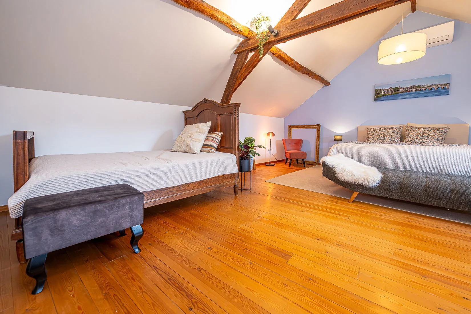 Een sfeervol slaapkamer met houten vloer, een bedstee met witte beddengoed en een groot bed met gestreept beddengoed, een plafond met zichtbare houten balken, een muur met een schilderij van een rivier en brug, planten, lampen en decoratieve elemente