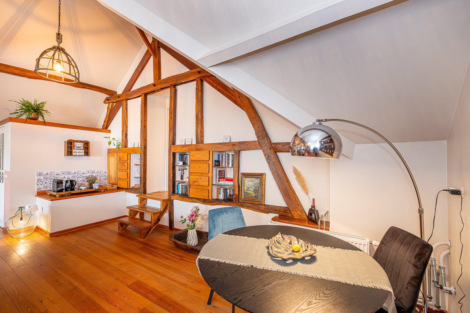 Interieur van een woonkamer met een houten vloer en houten dwarsbalken, een ronde tafel met een beige tafelkleed en een decoratief dienblad, een paar eetkamerstoelen, een boekenkast, lampen en decoratieve planten.